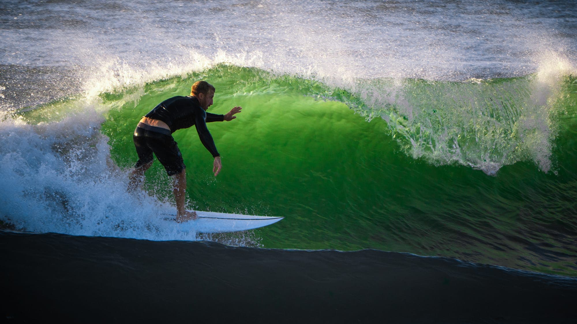 Surfer crouched low on a backlit emerald wave, spray curling overhead at golden hour