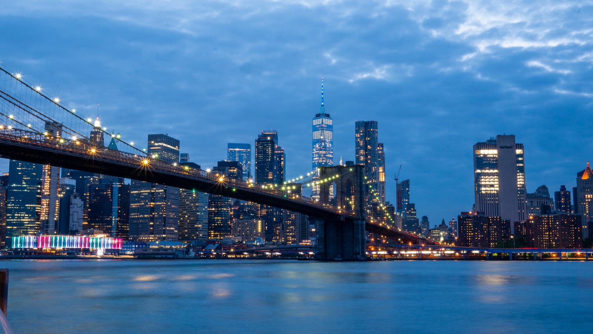 Brooklyn Bridge stretching toward the lit towers of Lower Manhattan at blue hour, city lights reflecting on the East River.