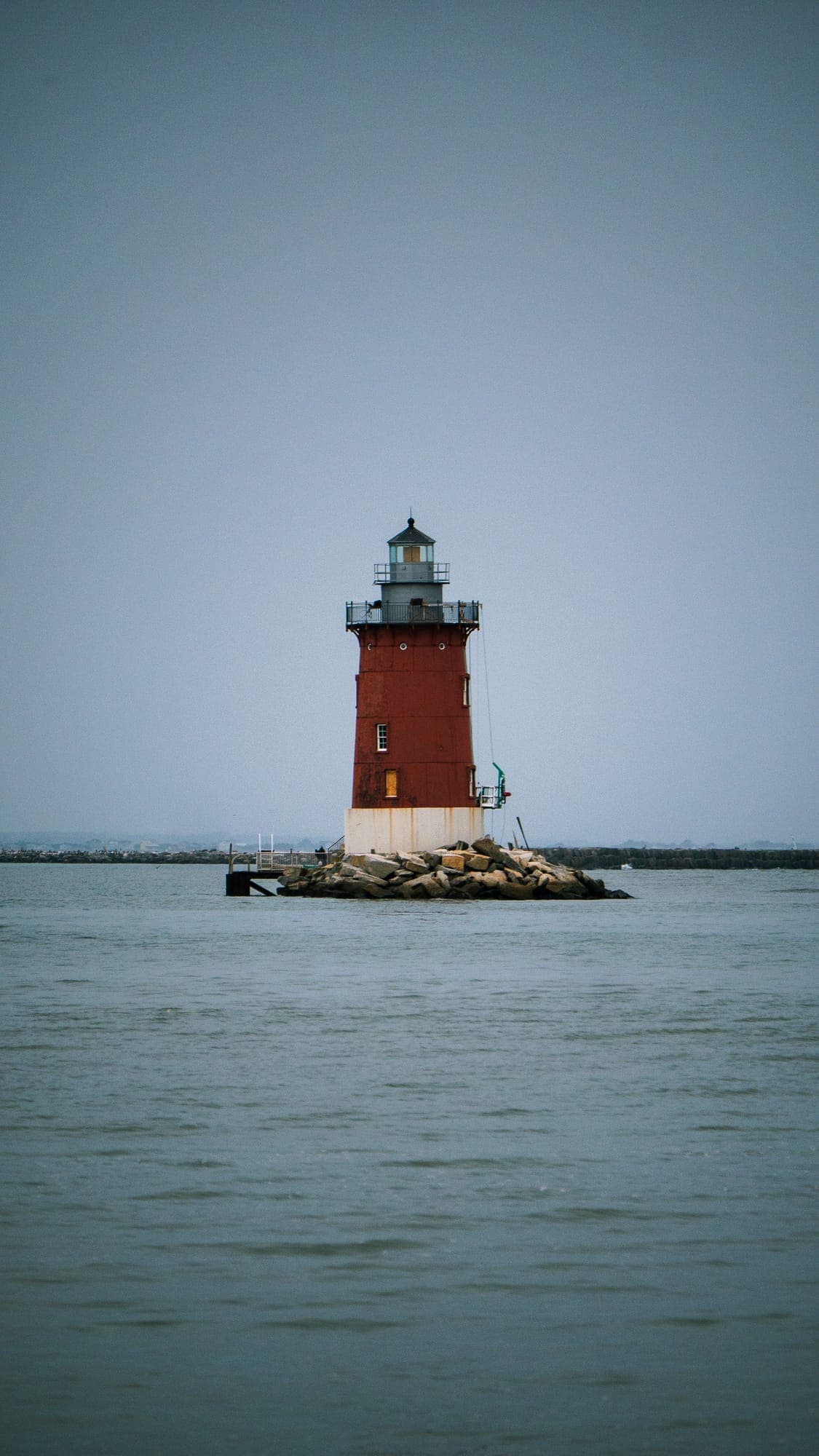 Red and white Delaware breakwater lighthouse standing alone on its rock pile in flat gray water beneath an empty sky.