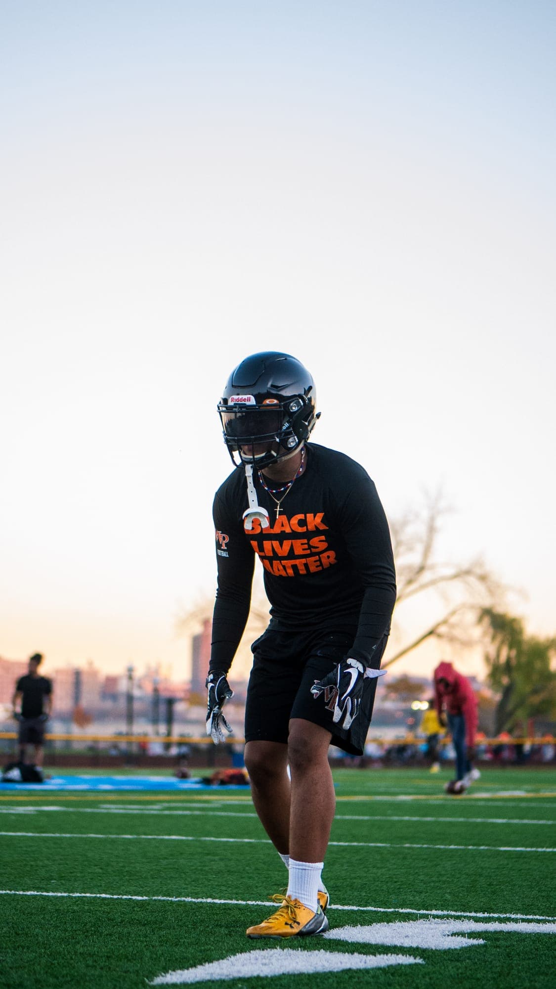 Helmeted football player in a Black Lives Matter shirt settling into a ready stance at evening practice