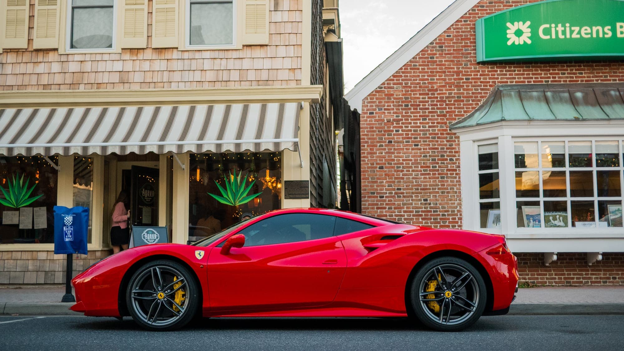 Red Ferrari 488 GTB in side profile in front of striped storefront awnings at dusk.