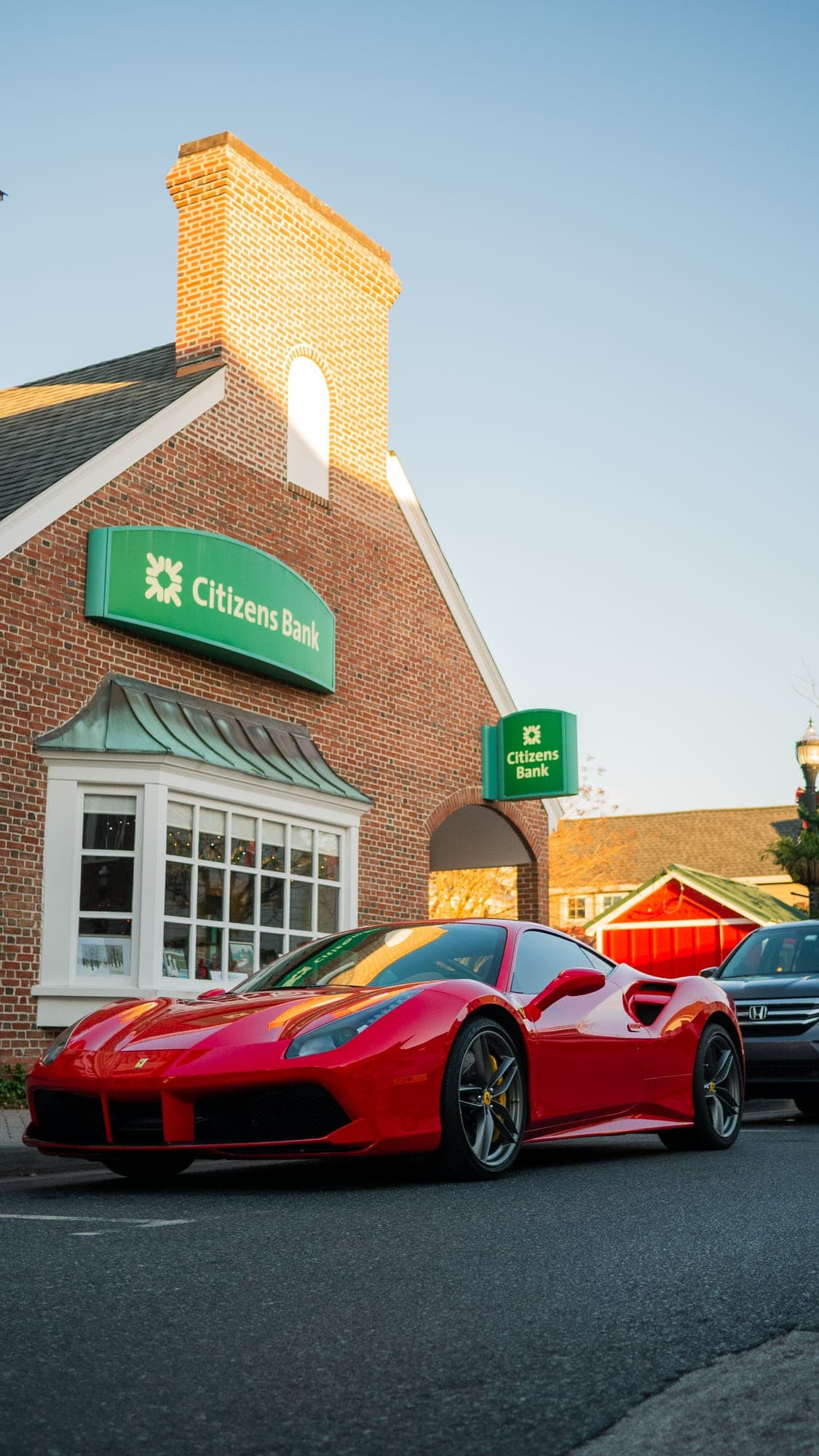 Low front three-quarter of a red Ferrari 488 GTB outside a brick bank corner in golden light.