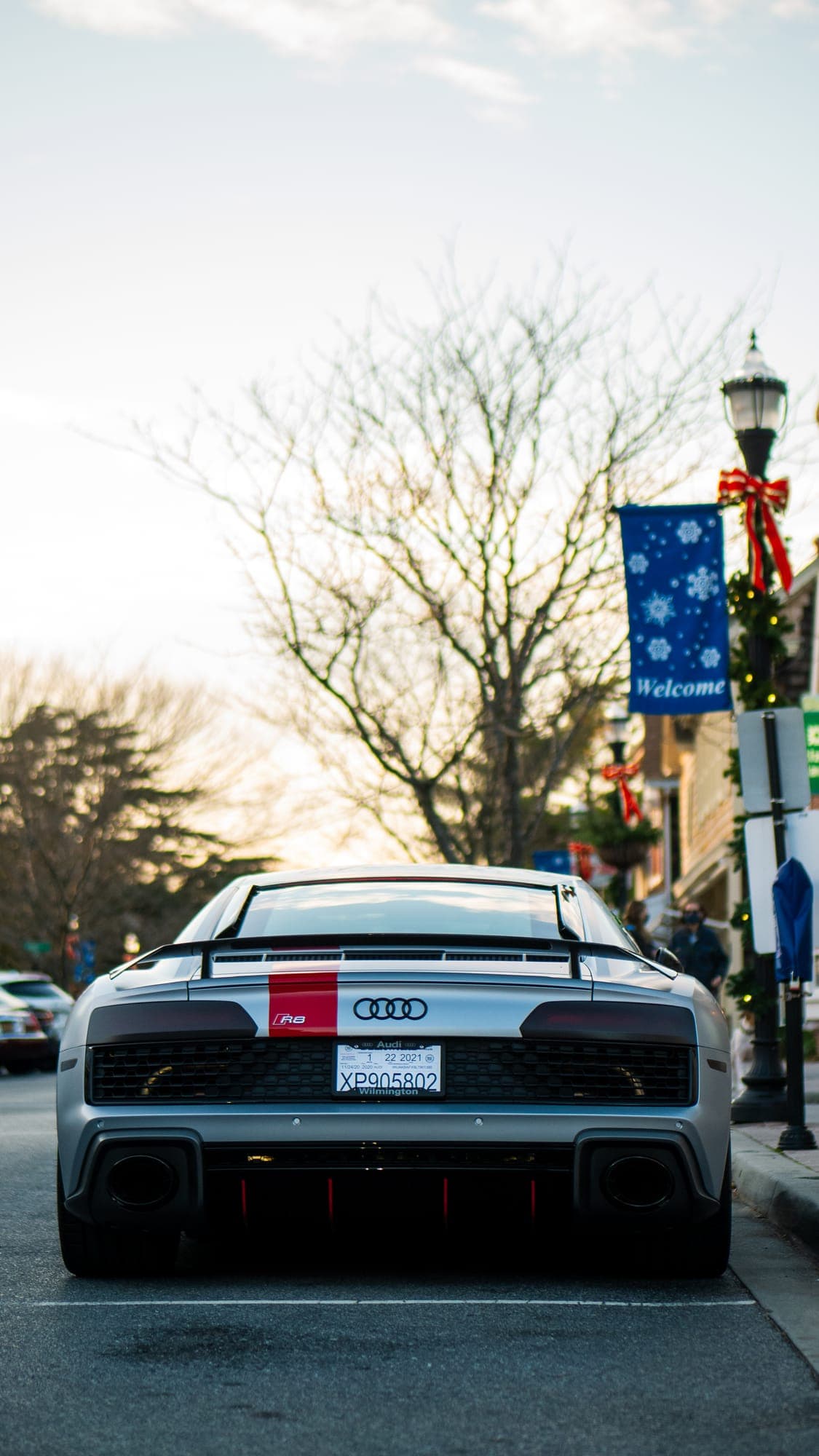 Silver Audi R8 with a red center stripe seen dead-on from behind beneath a welcome banner.
