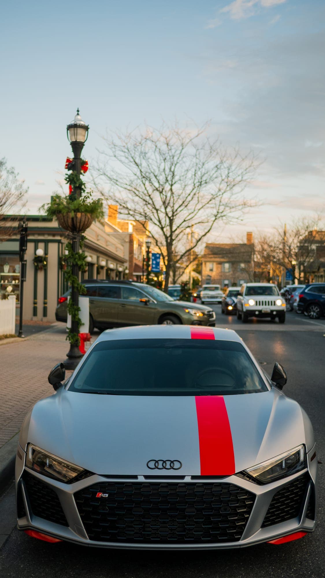 Head-on view of a silver Audi R8 with red stripe parked curbside as traffic passes behind.