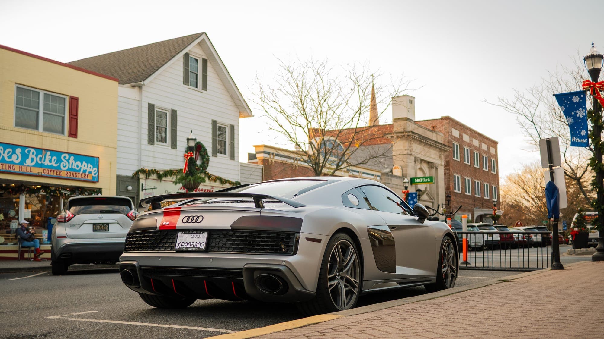 Rear view of a matte gray Audi R8 with red trim and wing, parked outside a bake shop on a festive main street.