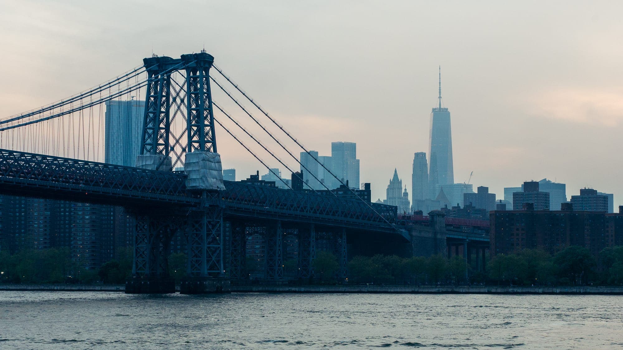 Williamsburg Bridge crossing the East River in pastel haze at dusk, One World Trade Center faint on the skyline behind.