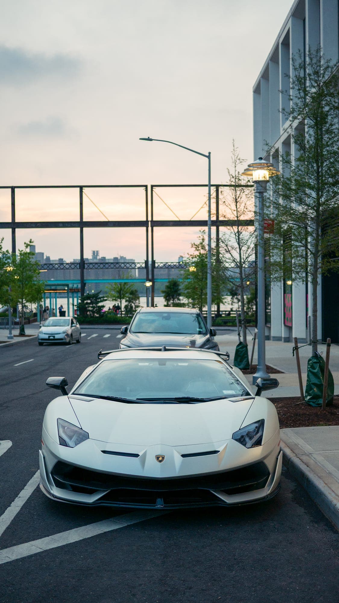 White Lamborghini Aventador SVJ head-on at dusk, parked under a streetlamp by the waterfront.