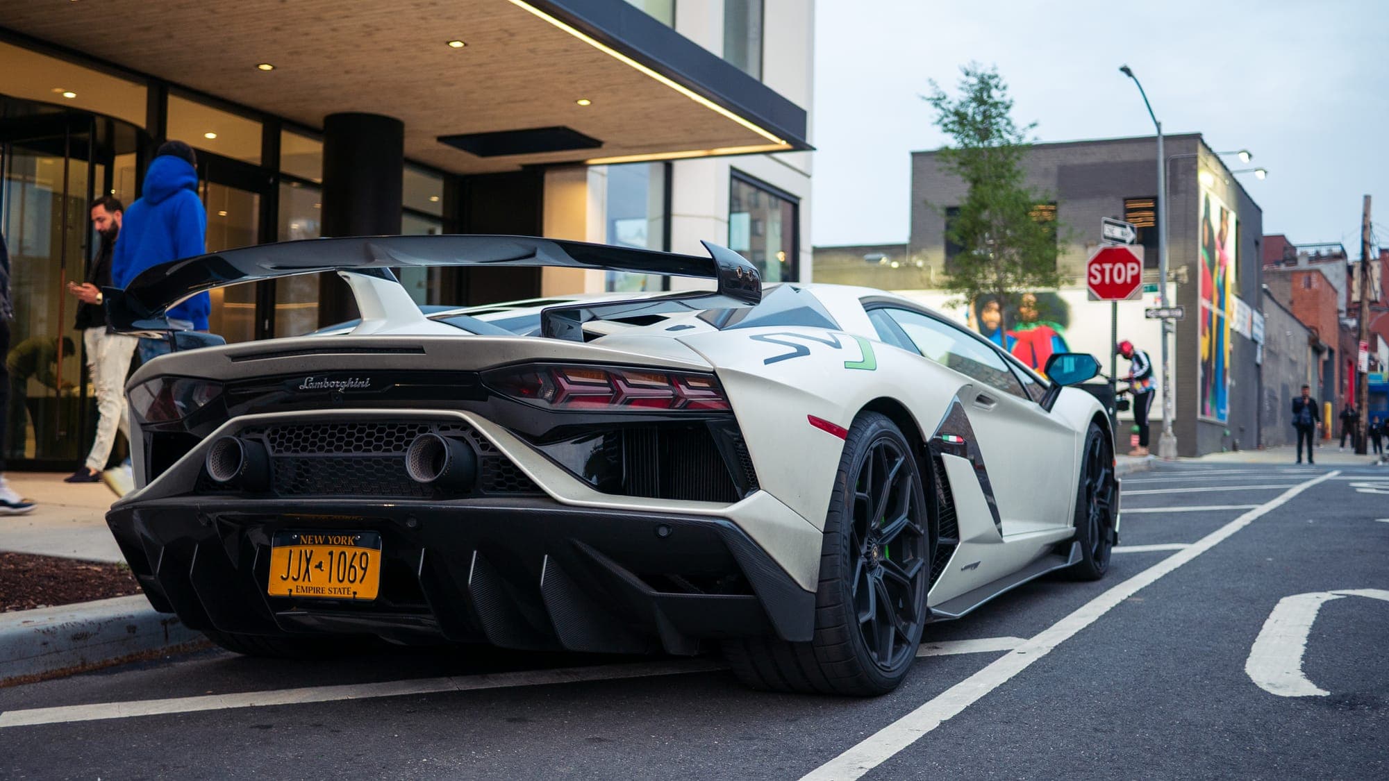 Rear three-quarter of a white Lamborghini Aventador SVJ with raised wing, curbside on a city street.