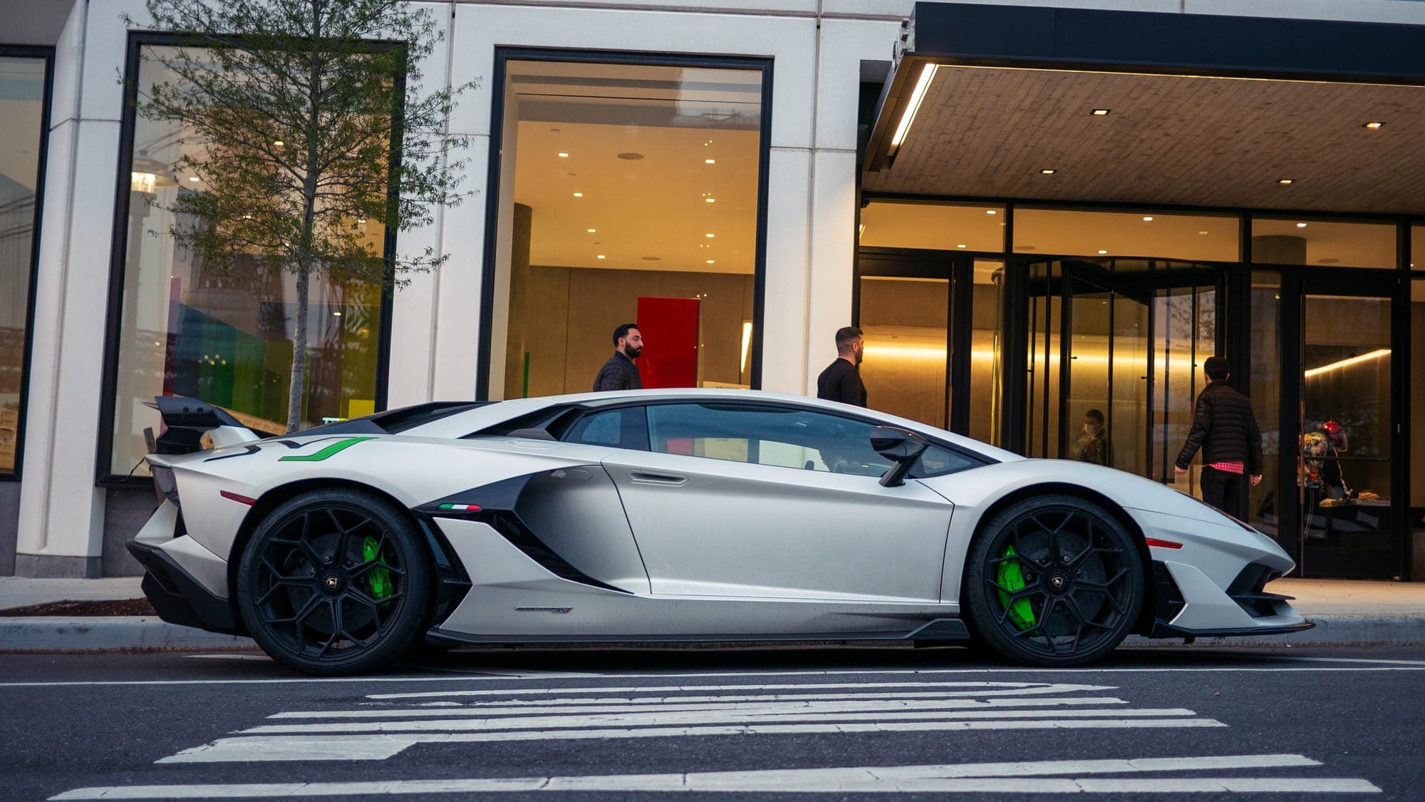 Side profile of a white Lamborghini Aventador SVJ with green brake calipers at a city crosswalk.