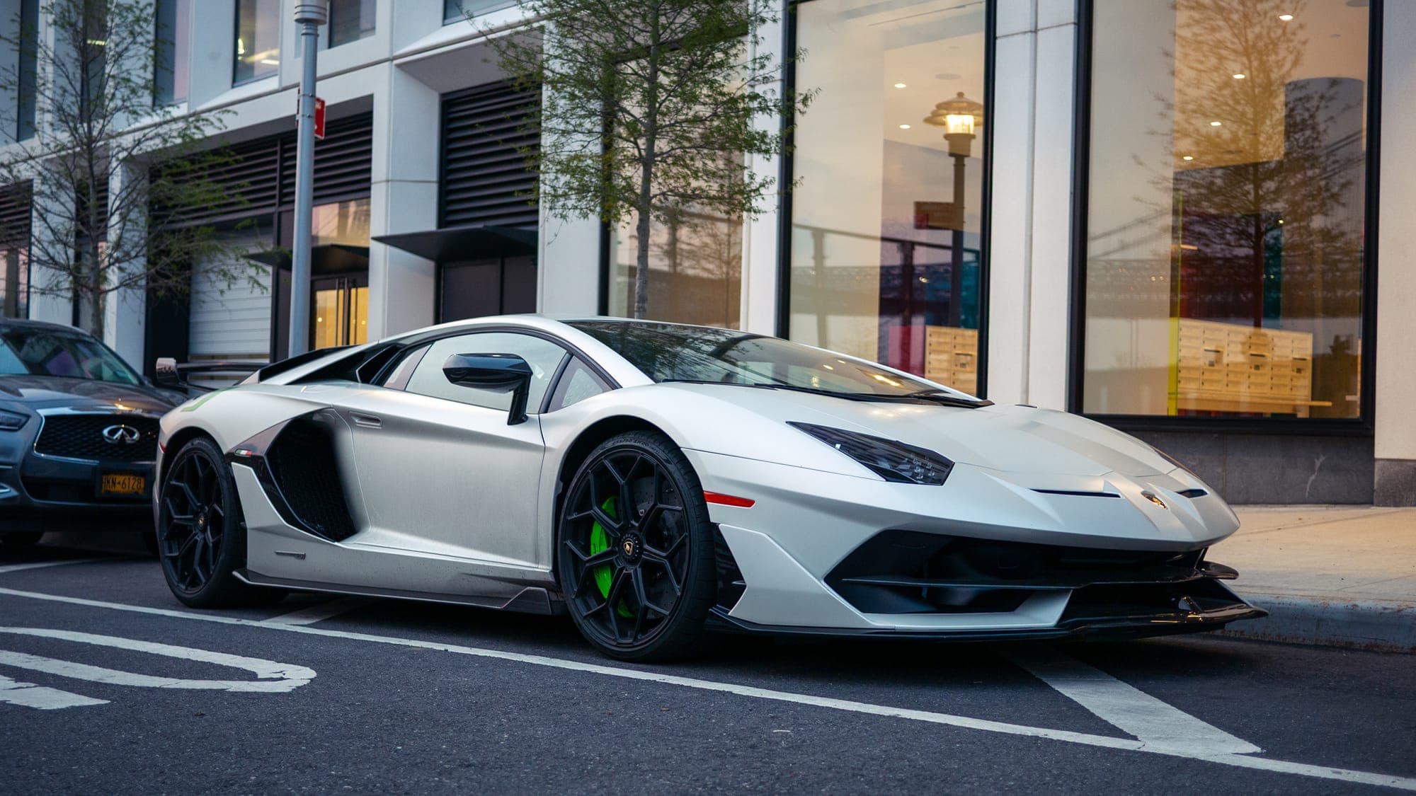 Front three-quarter of a white Lamborghini Aventador SVJ on black wheels, parked along a glass-fronted city block.