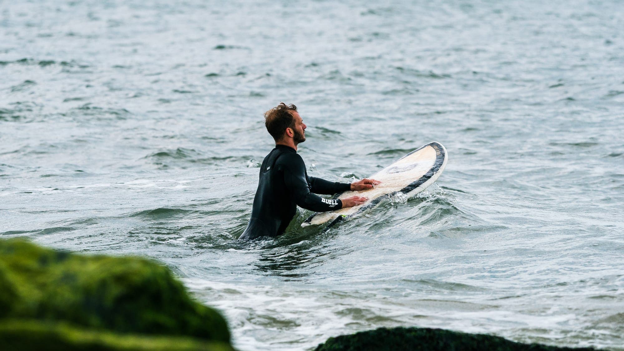 Wetsuited surfer wading into choppy gray water, guiding his board through the swell