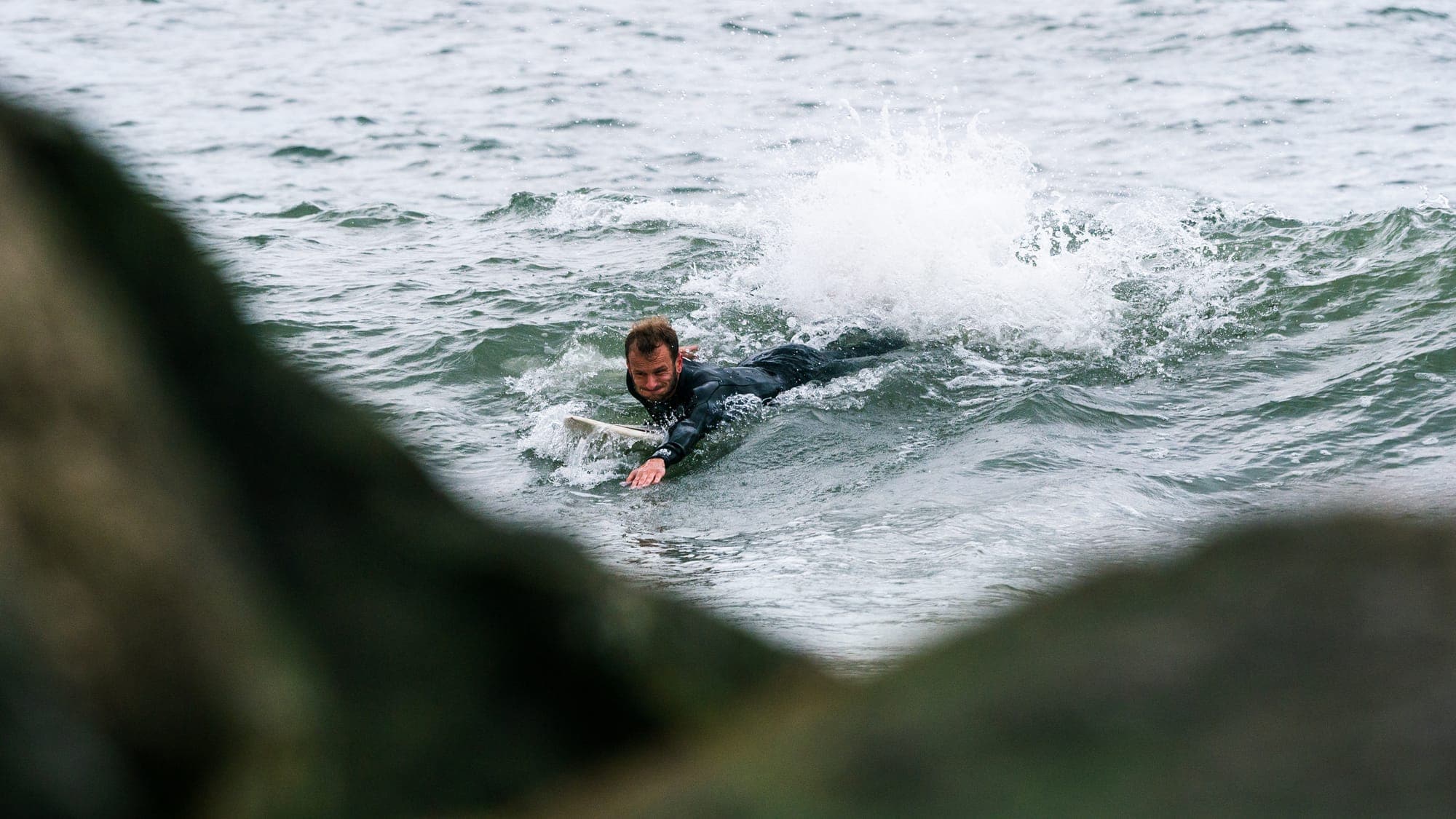 Surfer paddling hard into a breaking wave, framed between dark foreground rocks
