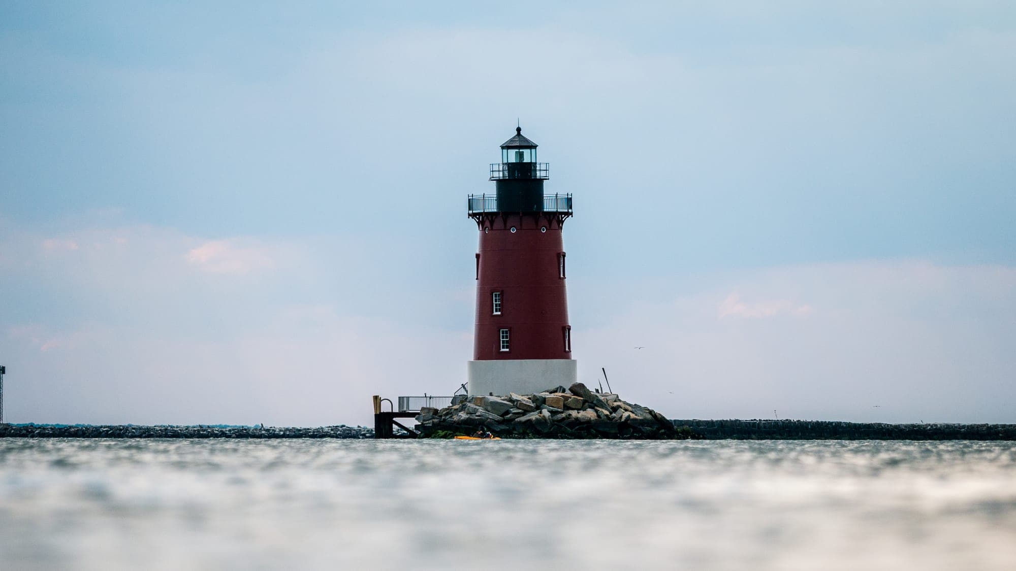 The red and white Delaware Breakwater lighthouse rises from a rocky jetty, seen from water level with a blurred wash of sea in the foreground under a cool evening sky.