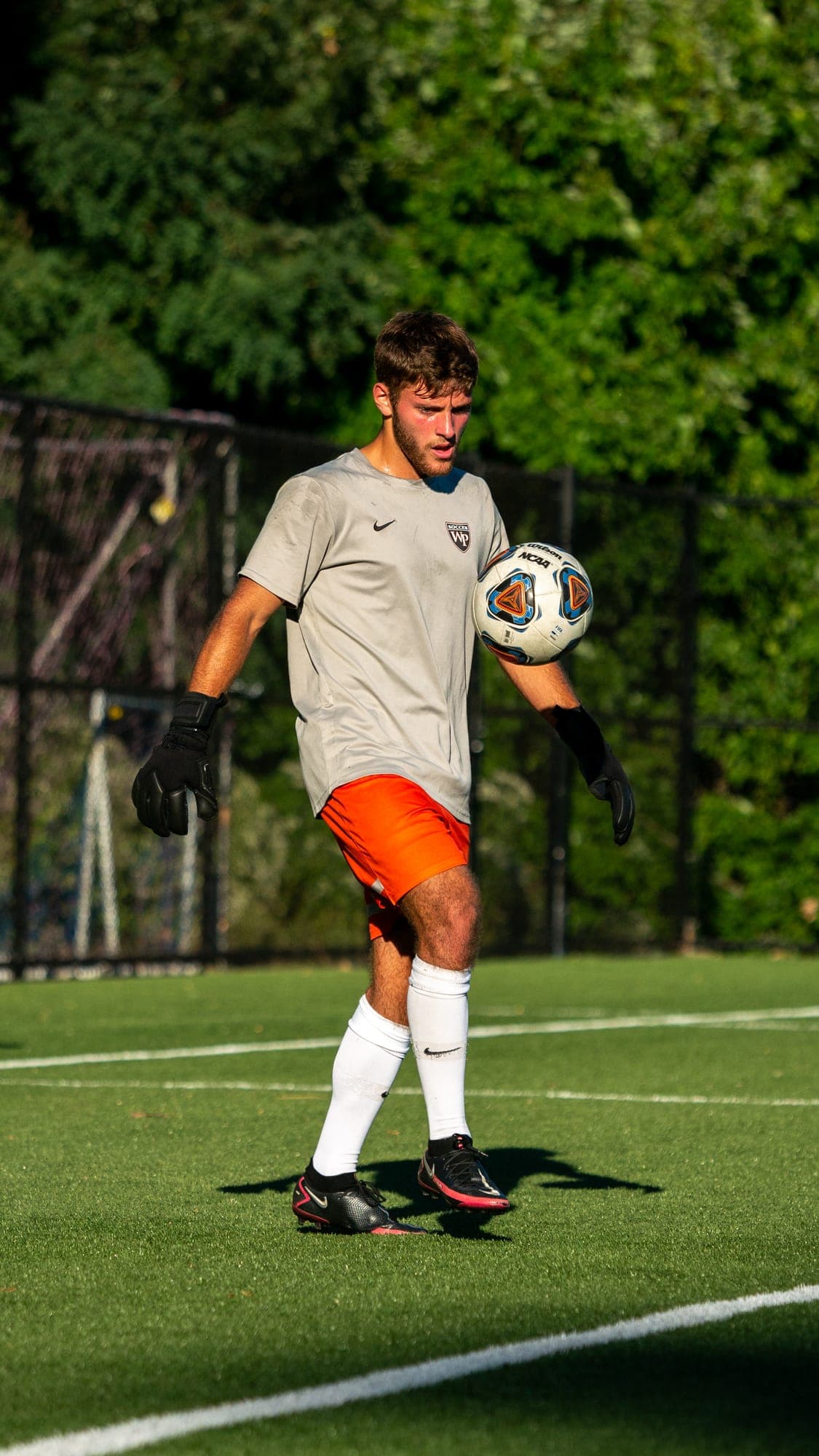 Goalkeeper in gloves and orange shorts juggling a ball off his thigh during sunlit training