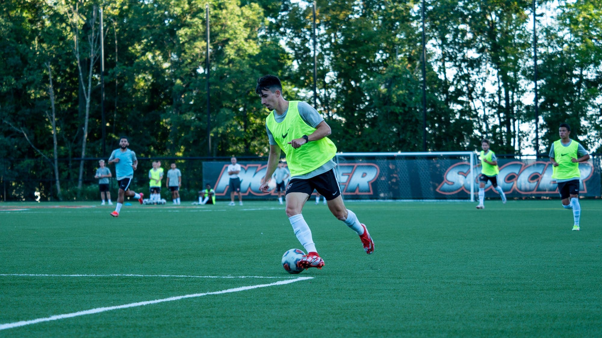 Soccer player in a neon bib dribbling upfield past banners, teammates spread across the pitch