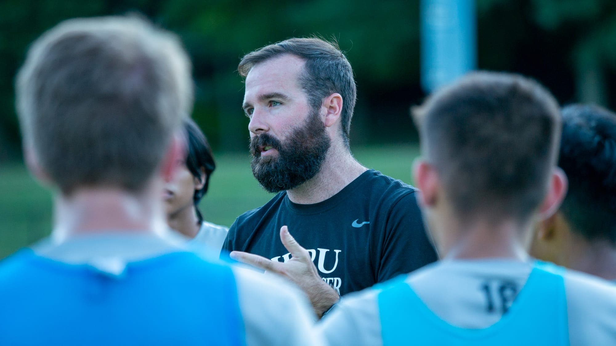Bearded coach gesturing mid-instruction inside a huddle of soccer players in blue bibs