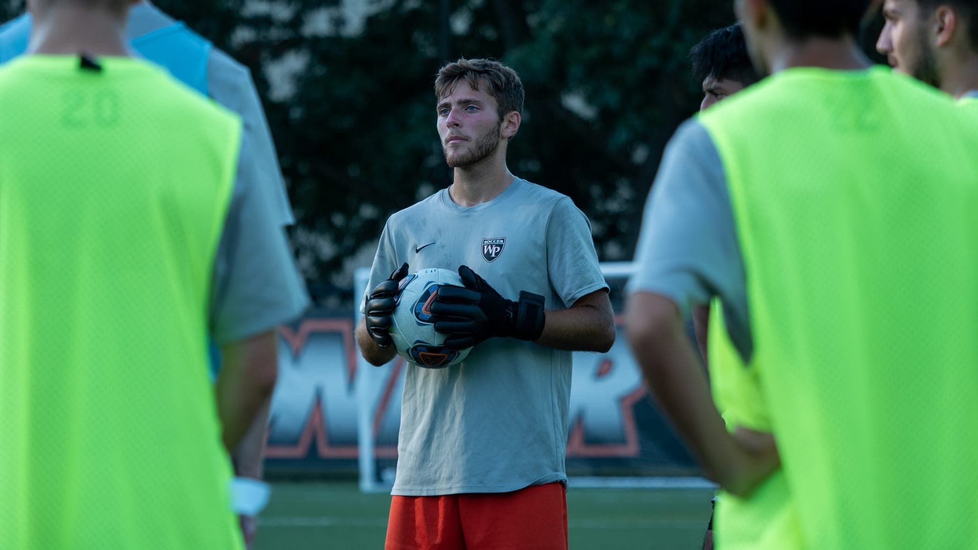 Goalkeeper in gloves clutching a soccer ball to his chest, listening in a team huddle at dusk
