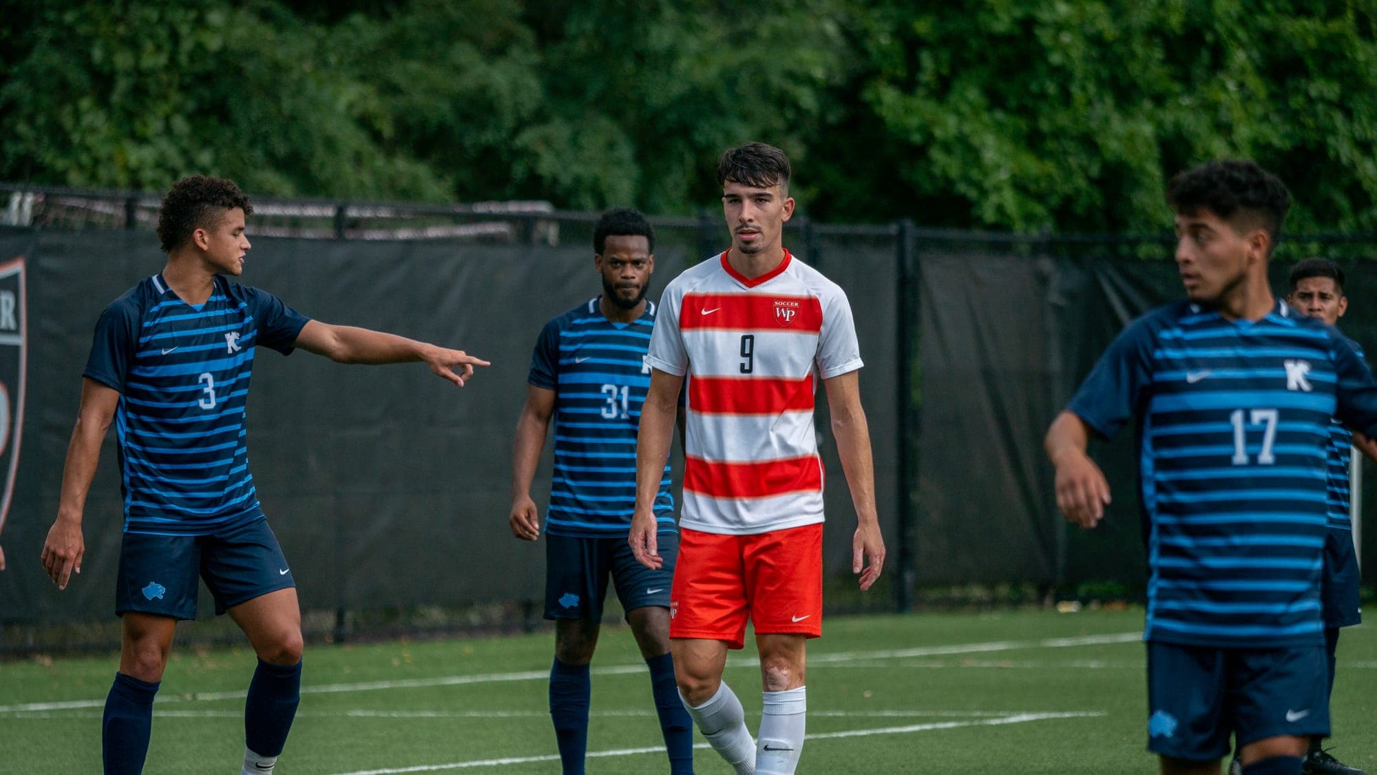 Soccer striker number 9 walking between blue-shirted Kean defenders as one points out a mark