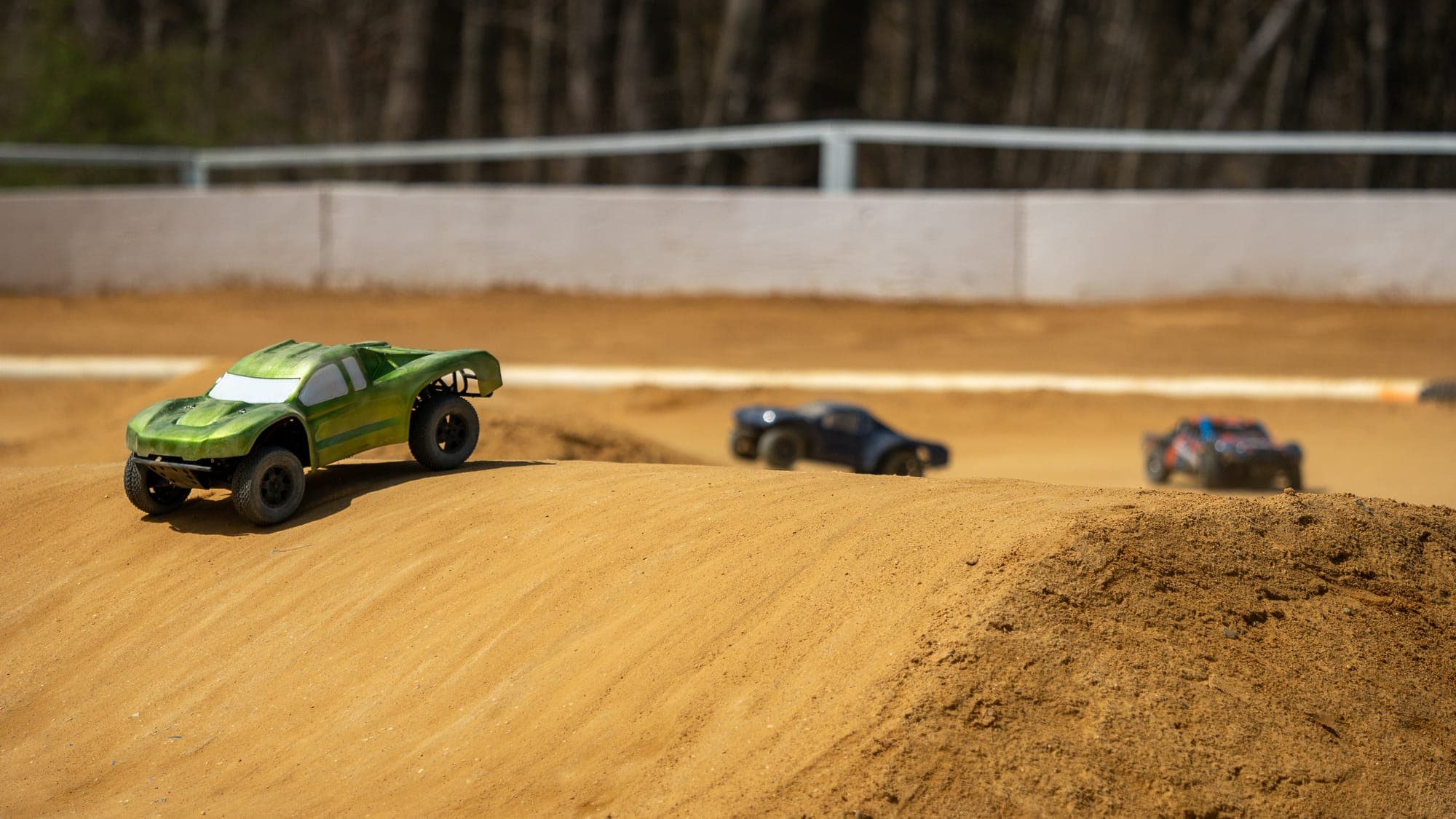 Green RC short-course truck cresting a dirt jump on an outdoor track, rivals blurred behind.