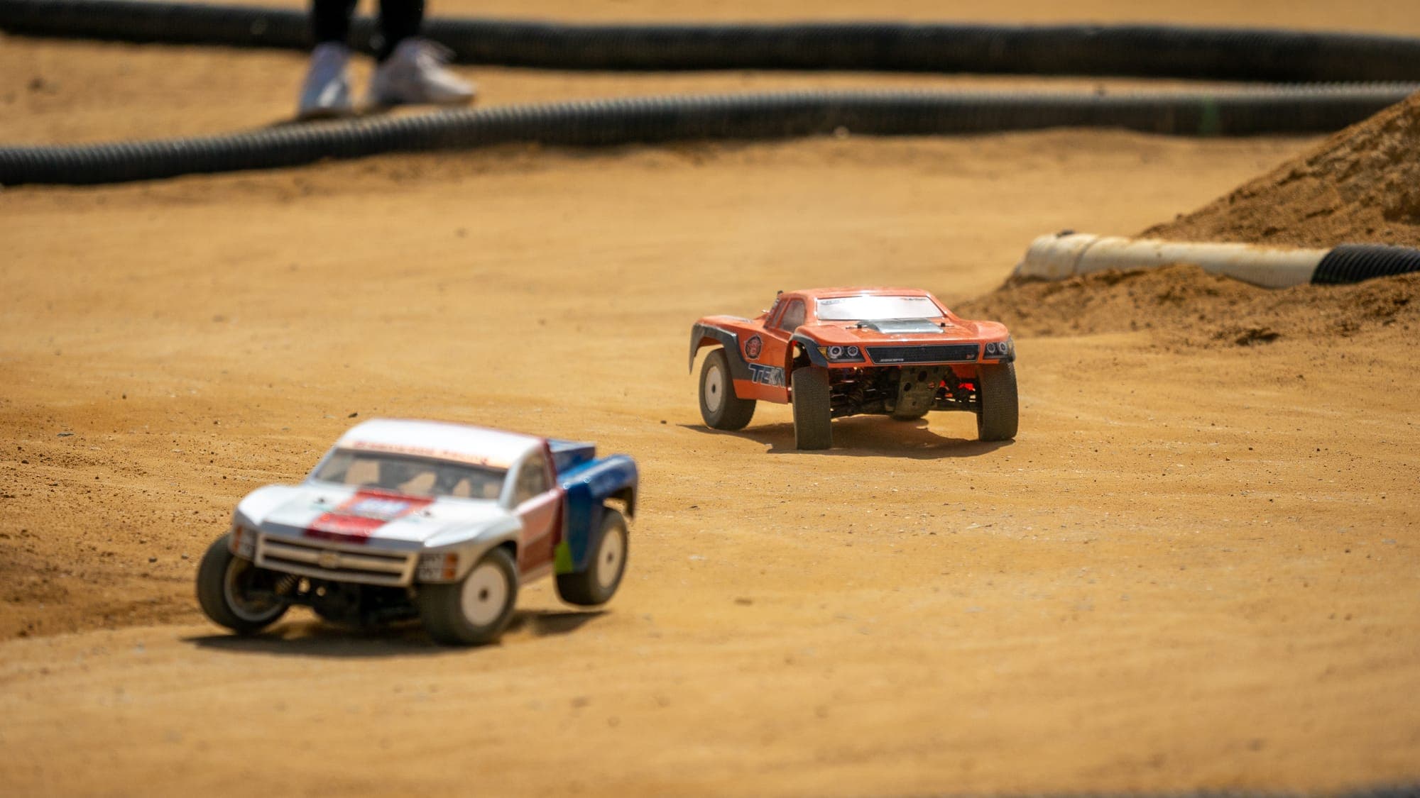Two RC short-course trucks racing through a corner on a dusty dirt track.