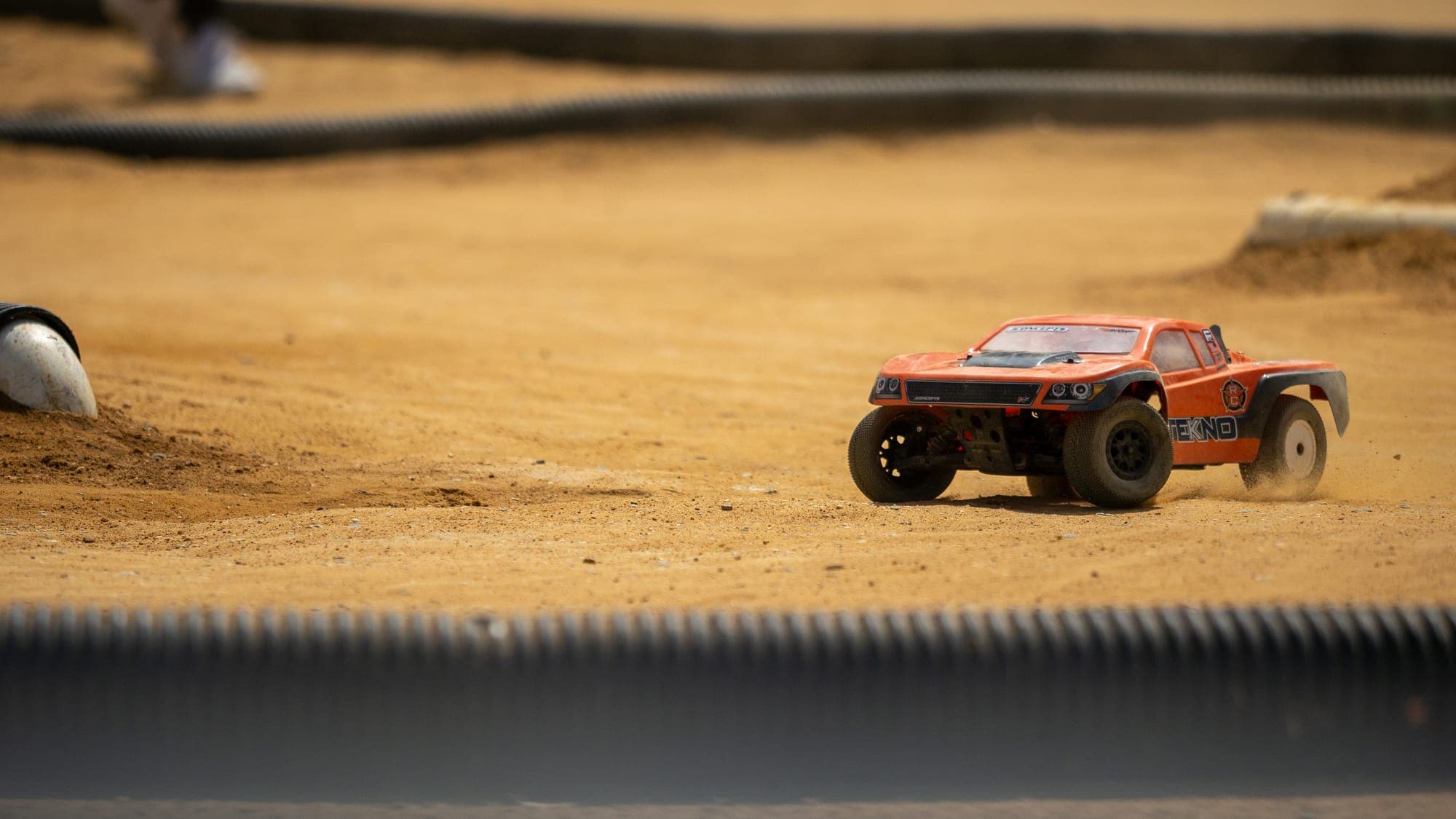 Orange Tekno RC truck kicking up dust mid-corner on a sunlit dirt track.