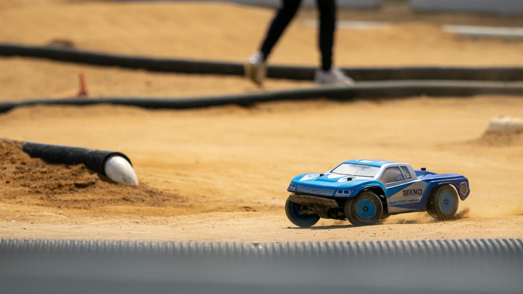 Blue Tekno RC truck sliding through a dusty corner, drainage-pipe barriers behind.