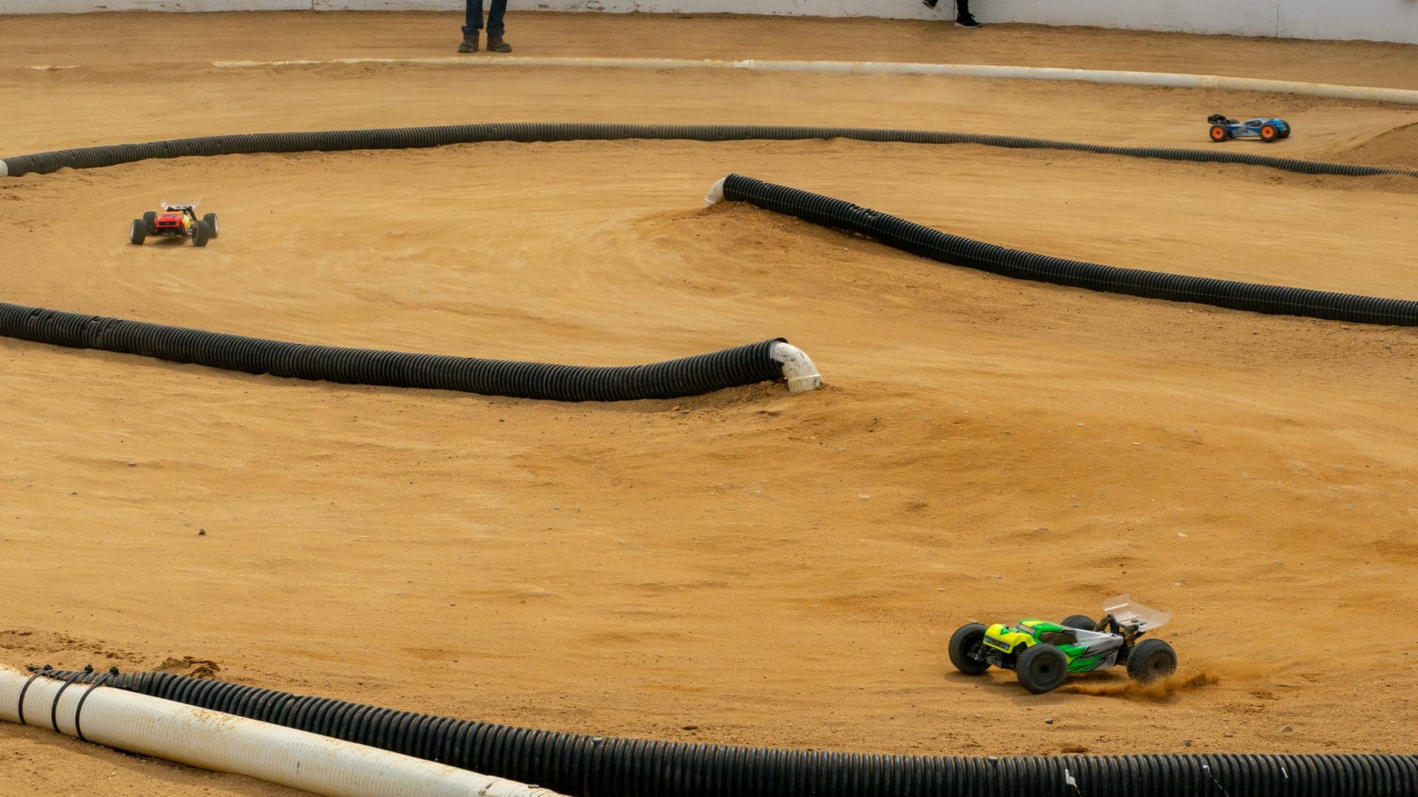 Wide view of an RC race in progress, three trucks spread across a banked dirt circuit.