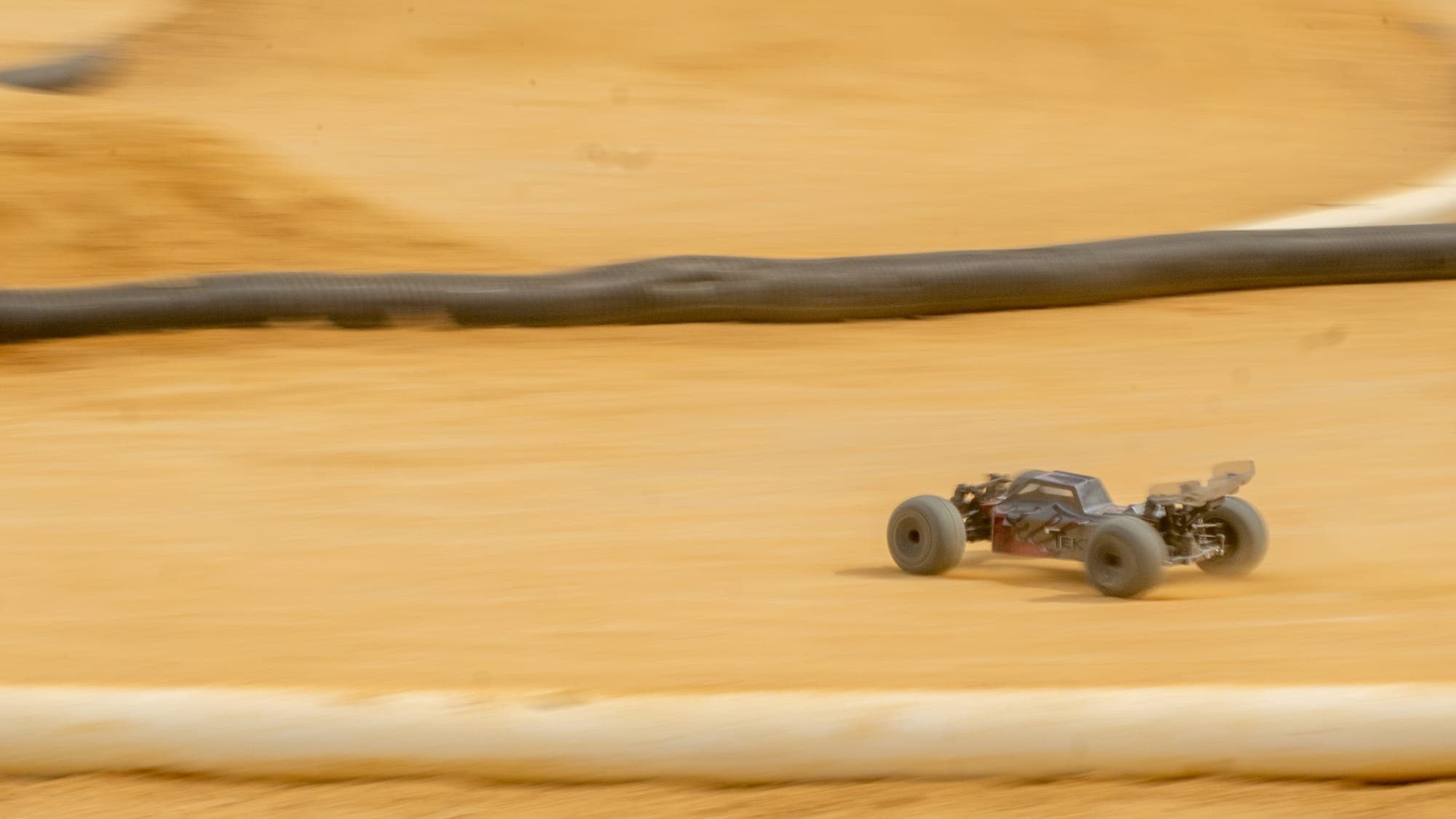 Tekno RC truggy speeding past a black track barrier on packed clay, heavy panning blur.