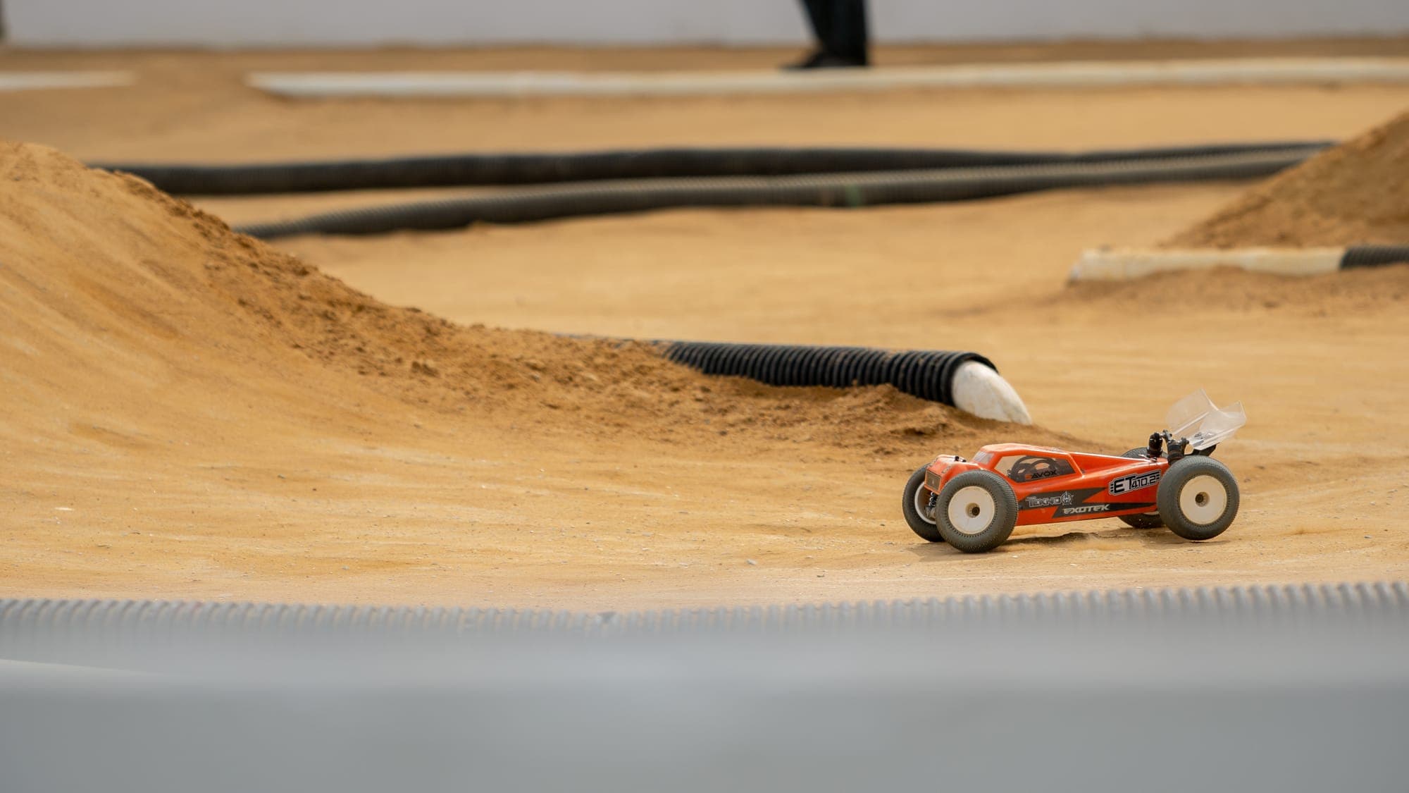Orange Tekno RC race buggy paused on a sculpted dirt berm of an indoor clay track.