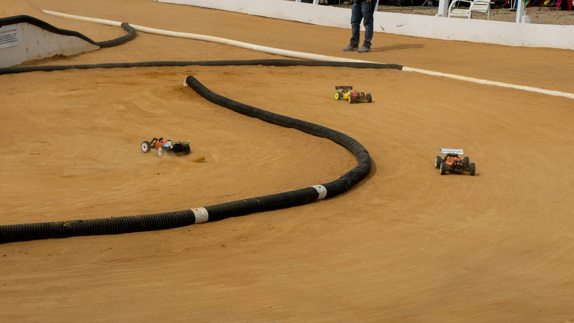 Three RC buggies spread across a sweeping dirt track corner lined with black hose barriers.