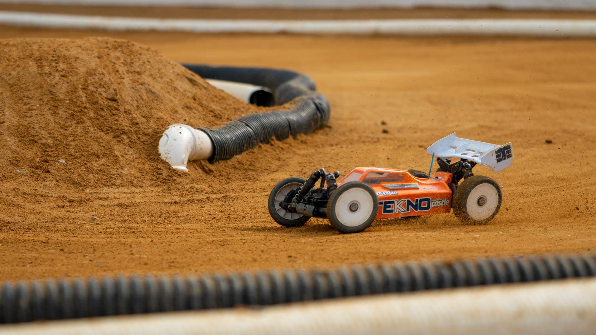 Orange-and-white Tekno RC buggy carving through loose dirt beside a drainage-pipe barrier.