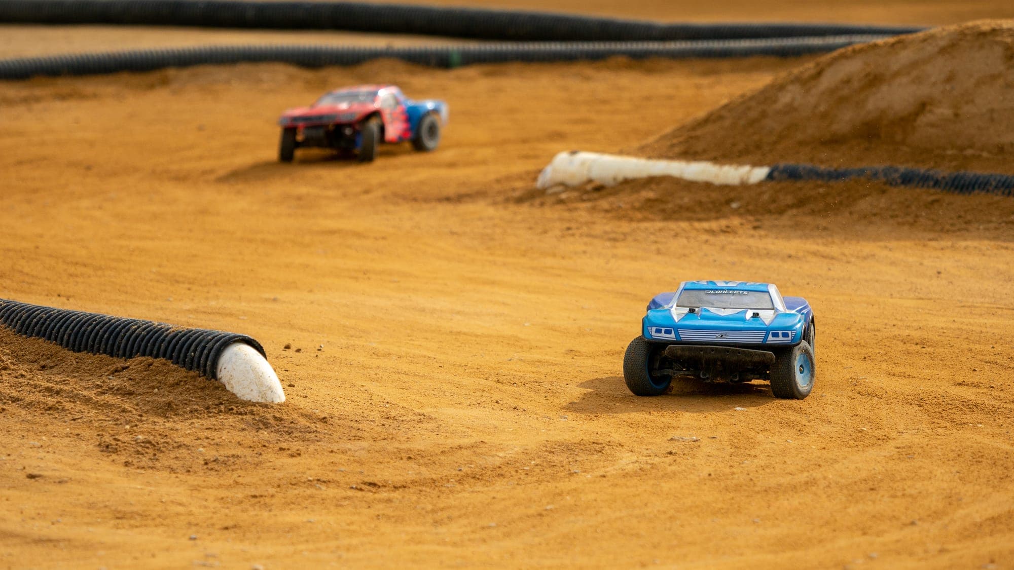 Blue JConcepts short-course RC truck leading a red rival through a dusty clay corner.
