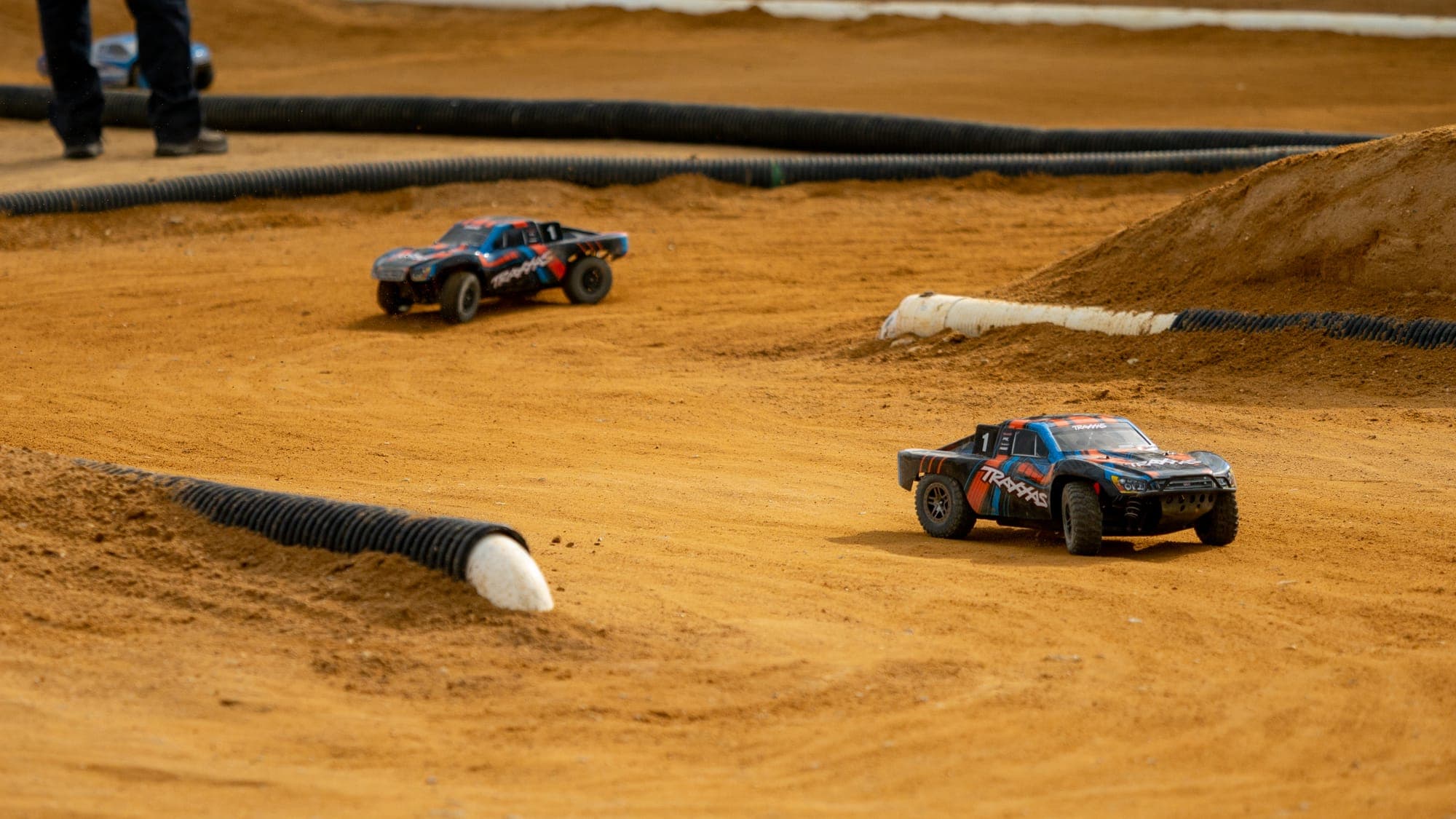 Two Traxxas short-course RC trucks racing nose to tail across rutted orange clay.