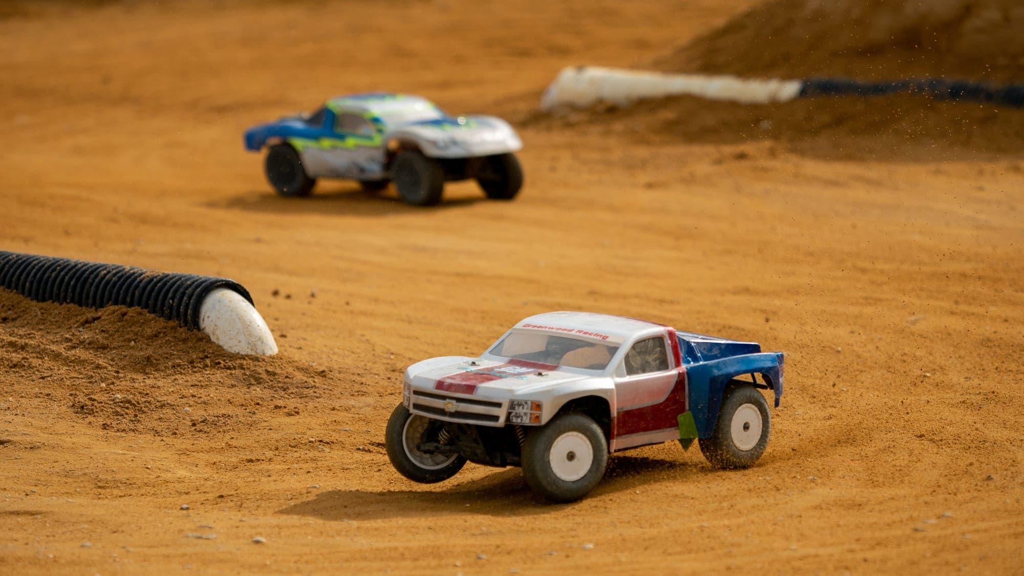 White, red, and blue short-course RC truck throwing dust off its rear wheel while a second truck trails behind.