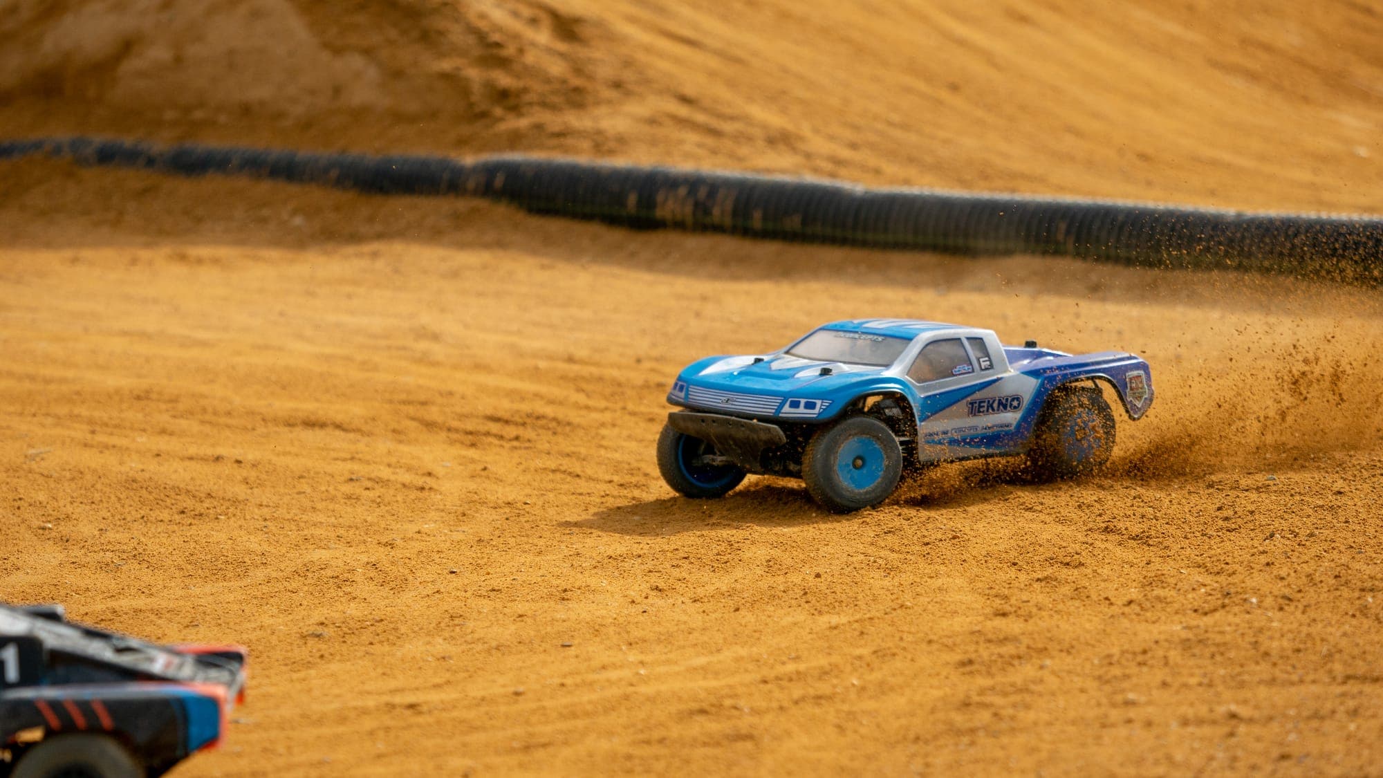 Blue Tekno short-course RC truck kicking up a rooster tail of dirt along a banked clay wall.