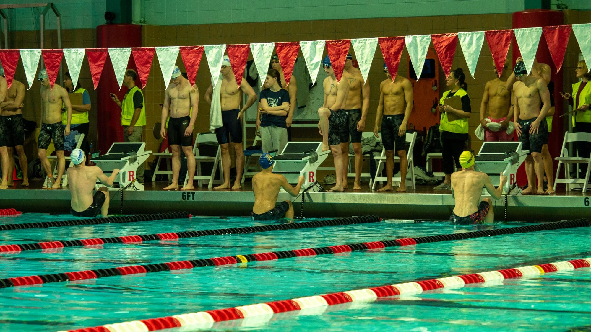 Swimmers lined up along the pool deck under red-and-white pennants, waiting behind the starting blocks