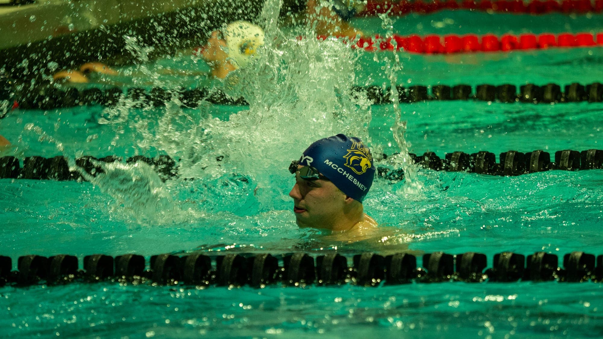 Swimmer in a blue cap breaking the surface in a burst of spray during a breaststroke race