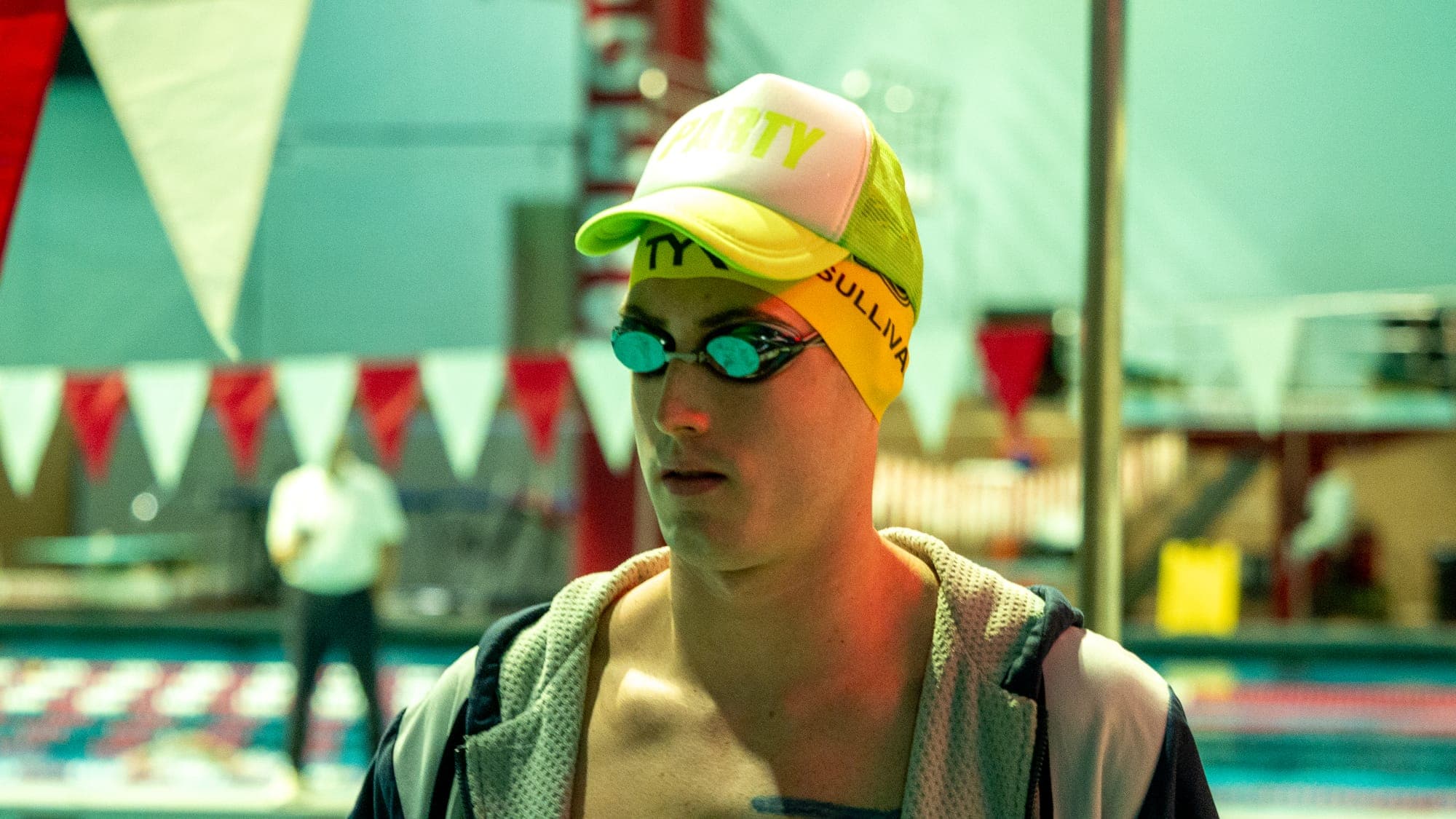 Swimmer in a yellow cap and mirrored goggles staring ahead before his race, pennant flags blurred behind him