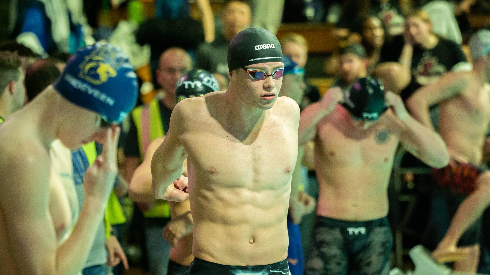 Shirtless swimmer in a black Arena cap striding through a crowded pool deck toward the starting blocks