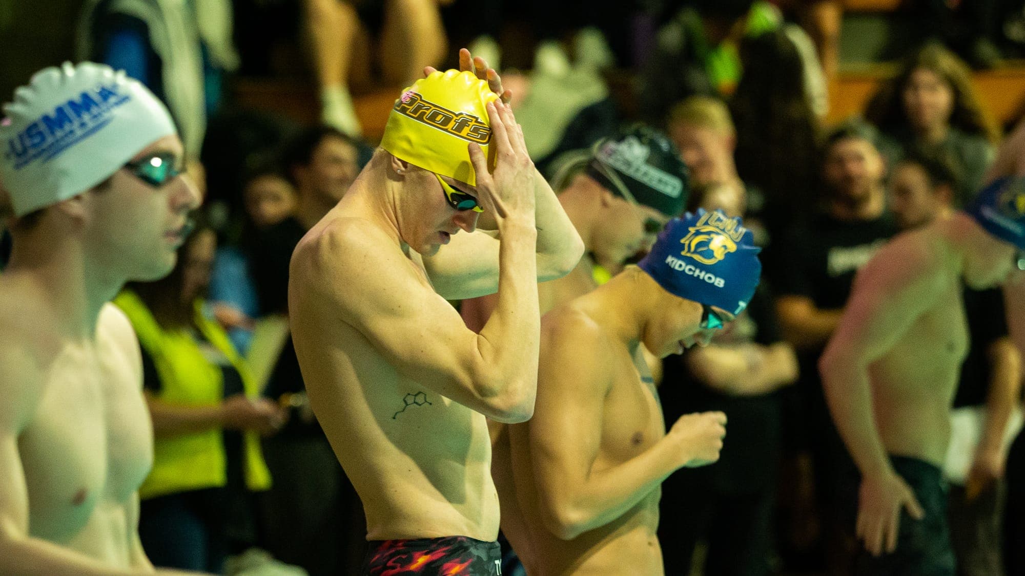 Swimmer pressing a yellow cap onto his head behind the blocks as competitors ready themselves around him