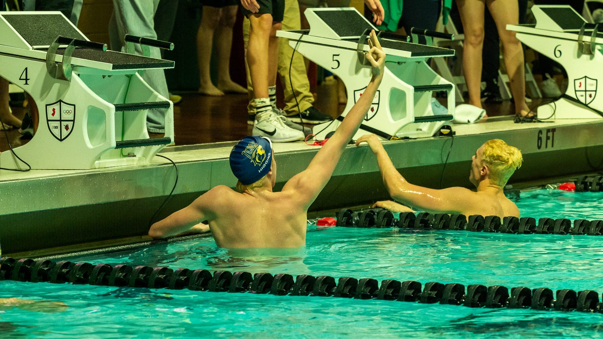 Swimmer reaching up from the water to slap hands with a teammate at the blocks after finishing a relay leg