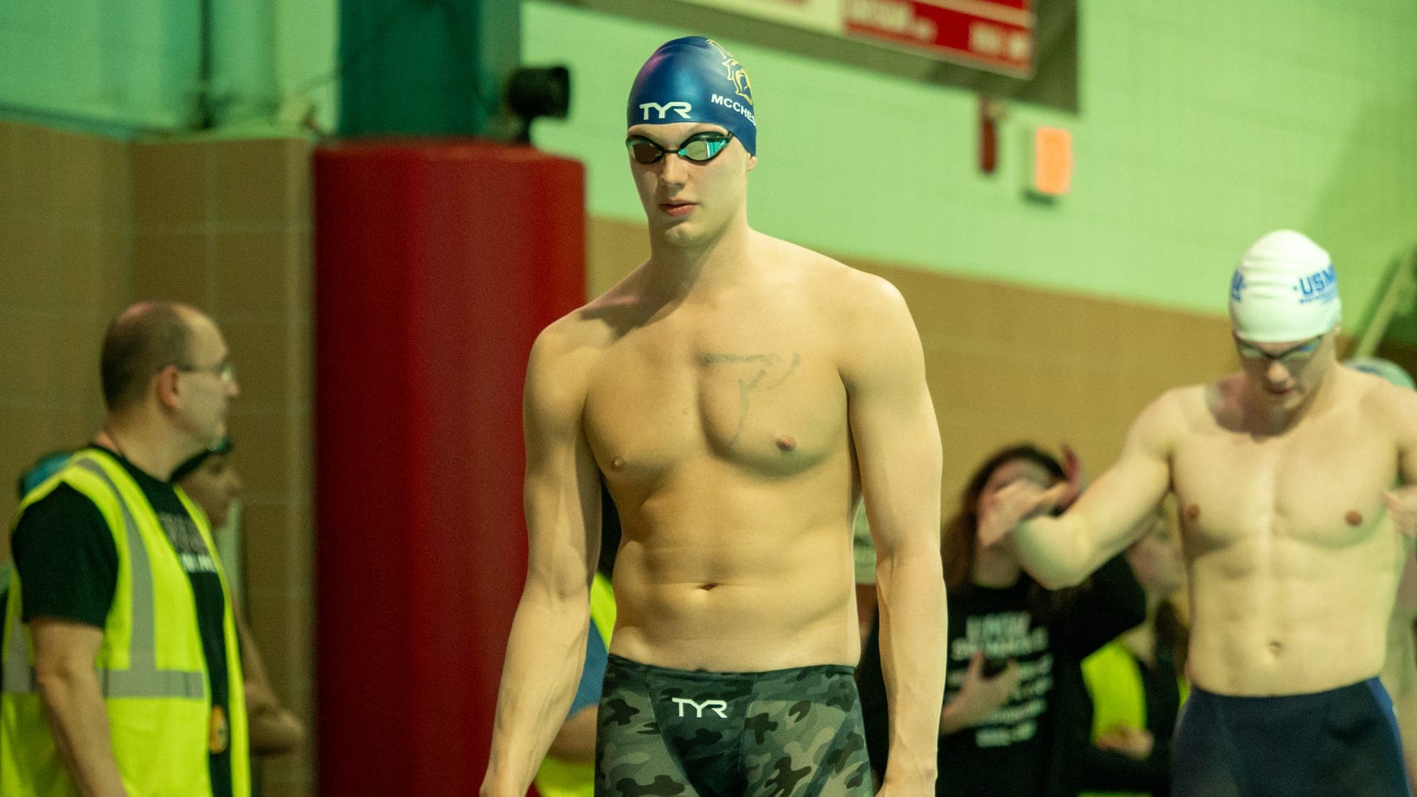 Swimmer in a camouflage racing suit walking the pool deck with a level stare, officials blurred behind him