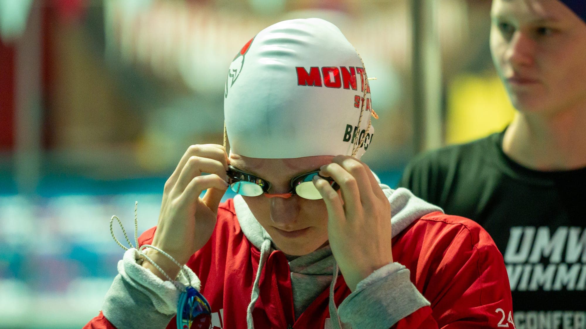 Swimmer in a red Montclair warm-up jacket seating her goggles beneath her cap before a race