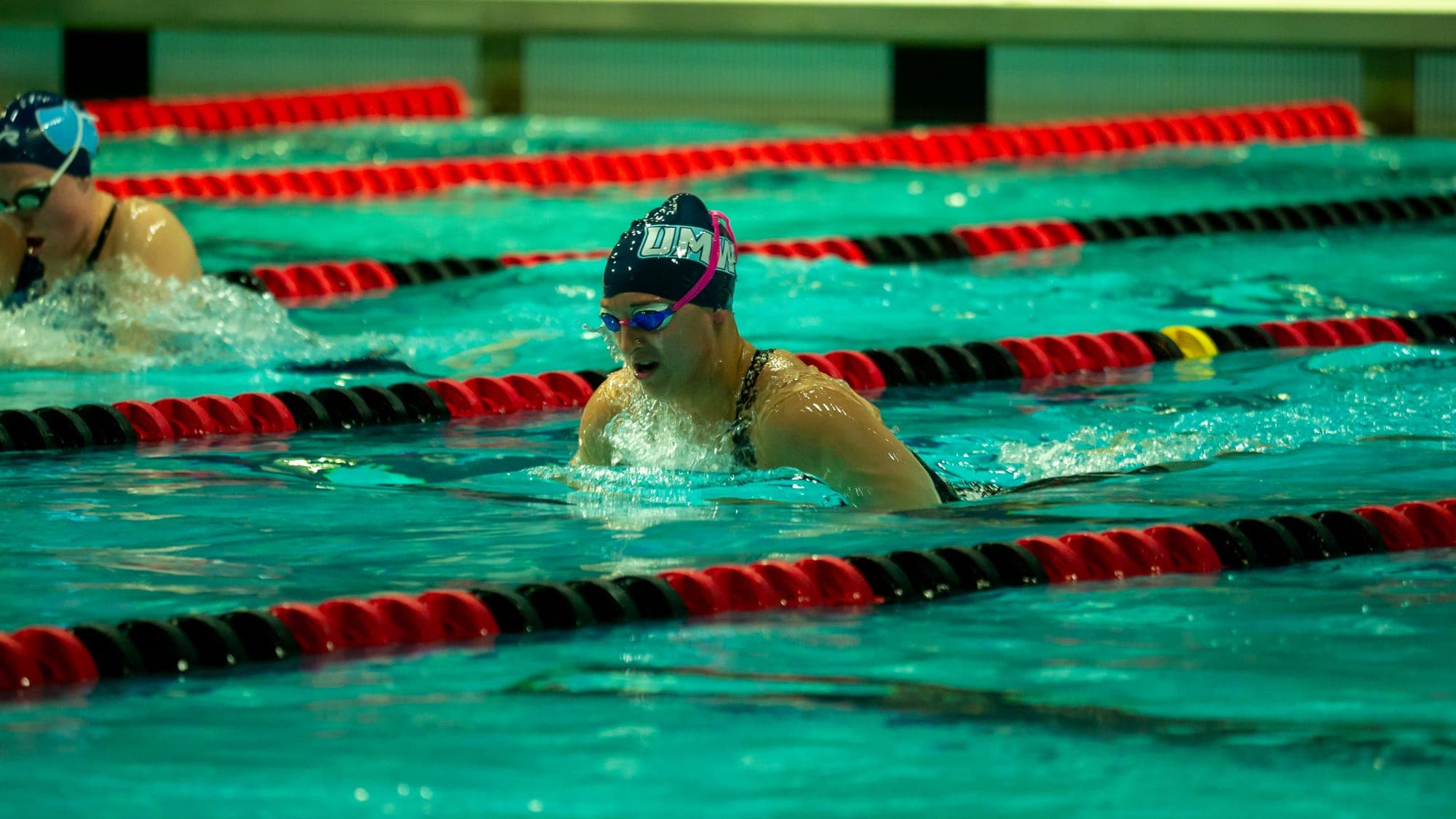 Breaststroker lifting her head for a breath between dark lane lines, a competitor surfacing in the next lane