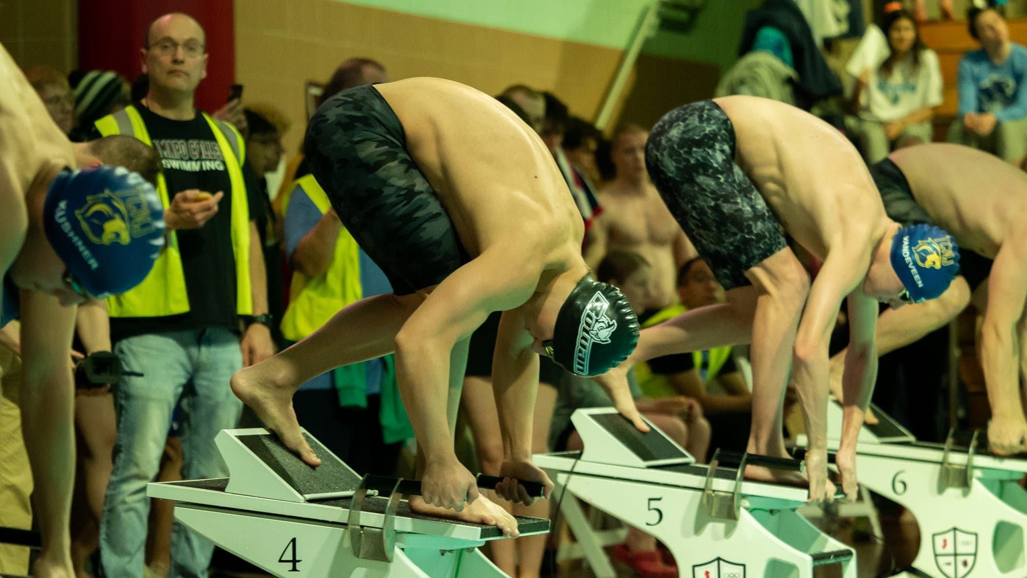 Swimmers coiled in the set position on starting blocks 4, 5 and 6, officials and spectators lining the pool deck behind them