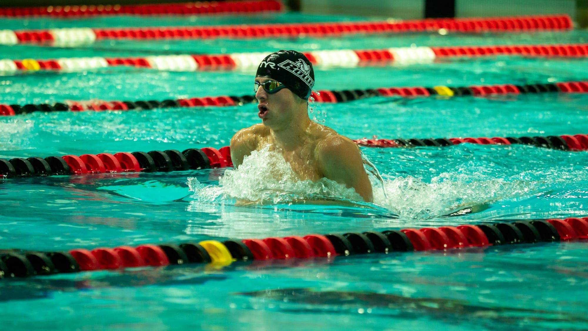 Breaststroke swimmer in a dark cap surging head-up out of turquoise water between red lane lines