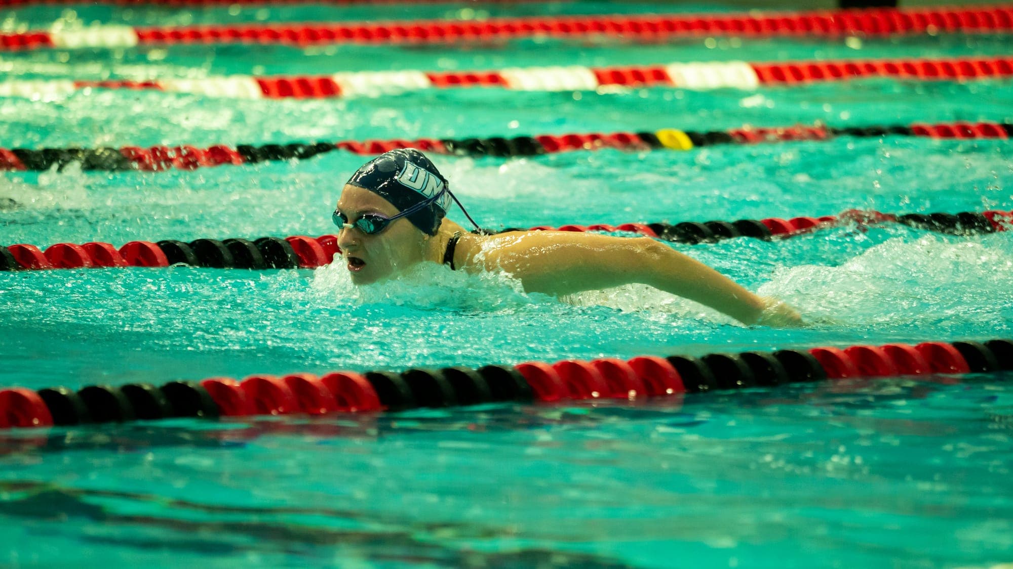 Female butterfly swimmer breaking the surface mid-stroke, arms sweeping back through the spray