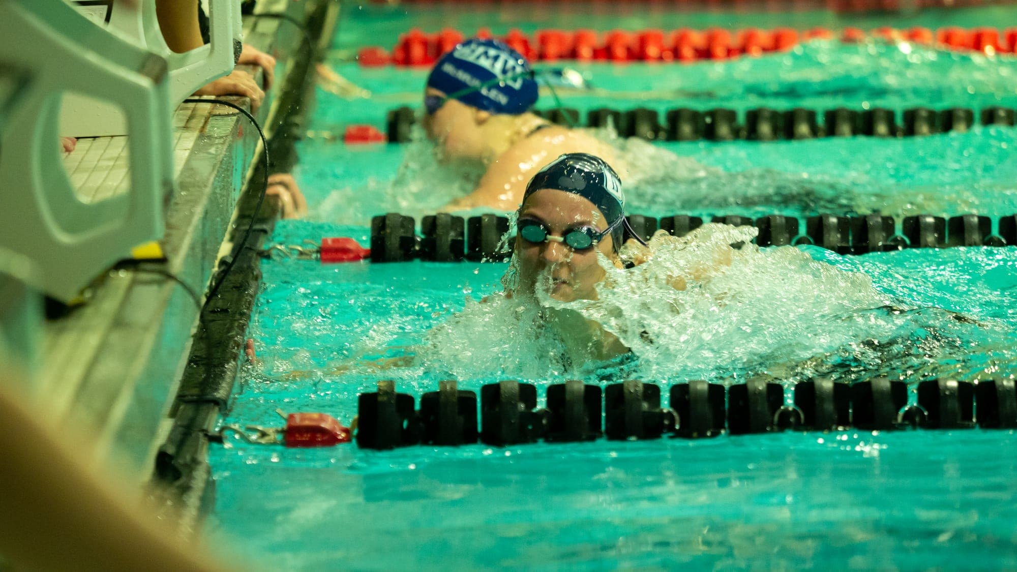 Female swimmer charging head-on through a burst of whitewater near the pool wall, another racer trailing behind
