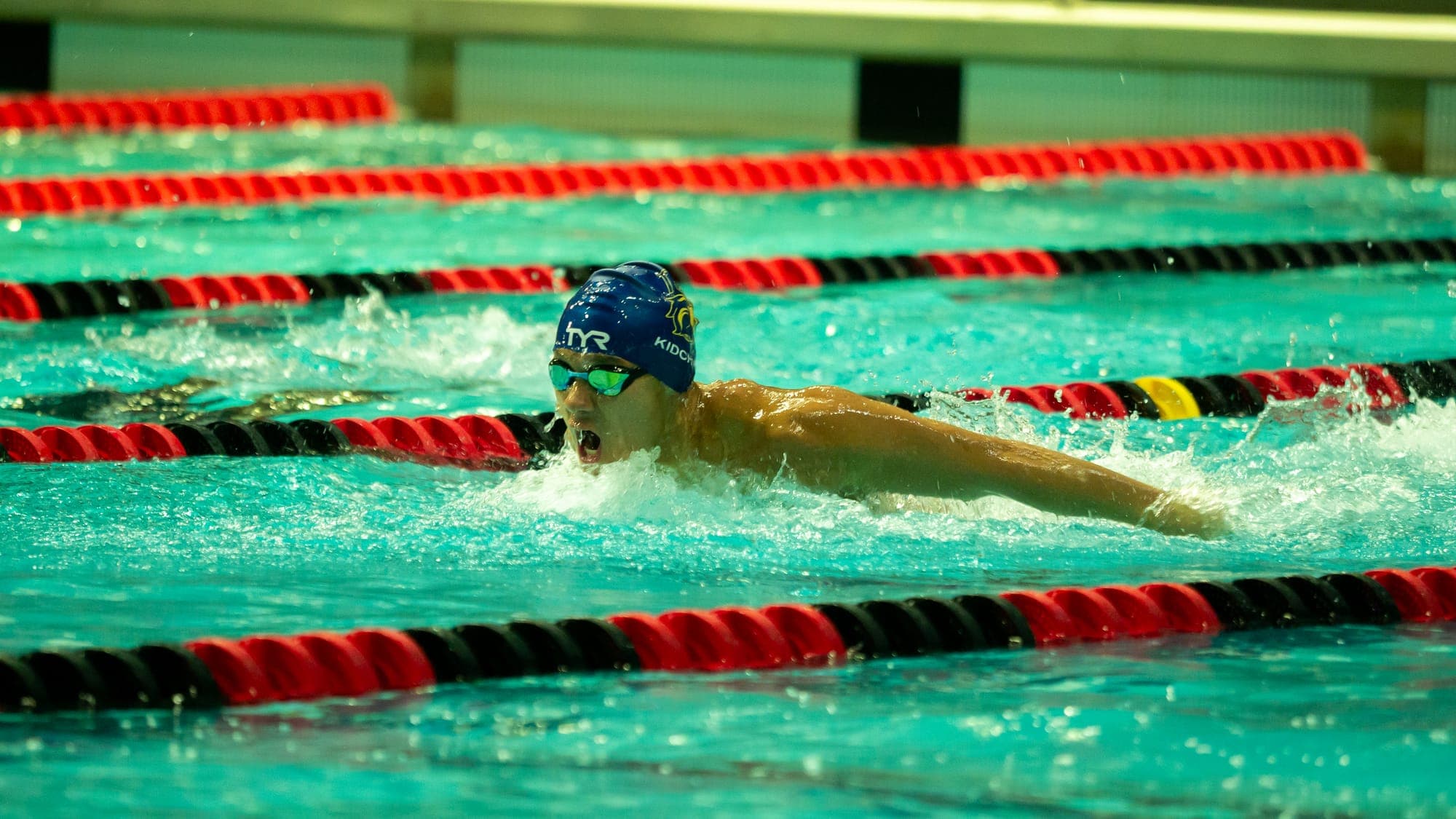 Butterfly swimmer in a blue cap mid-recovery, arms skimming the surface as spray trails off his hands