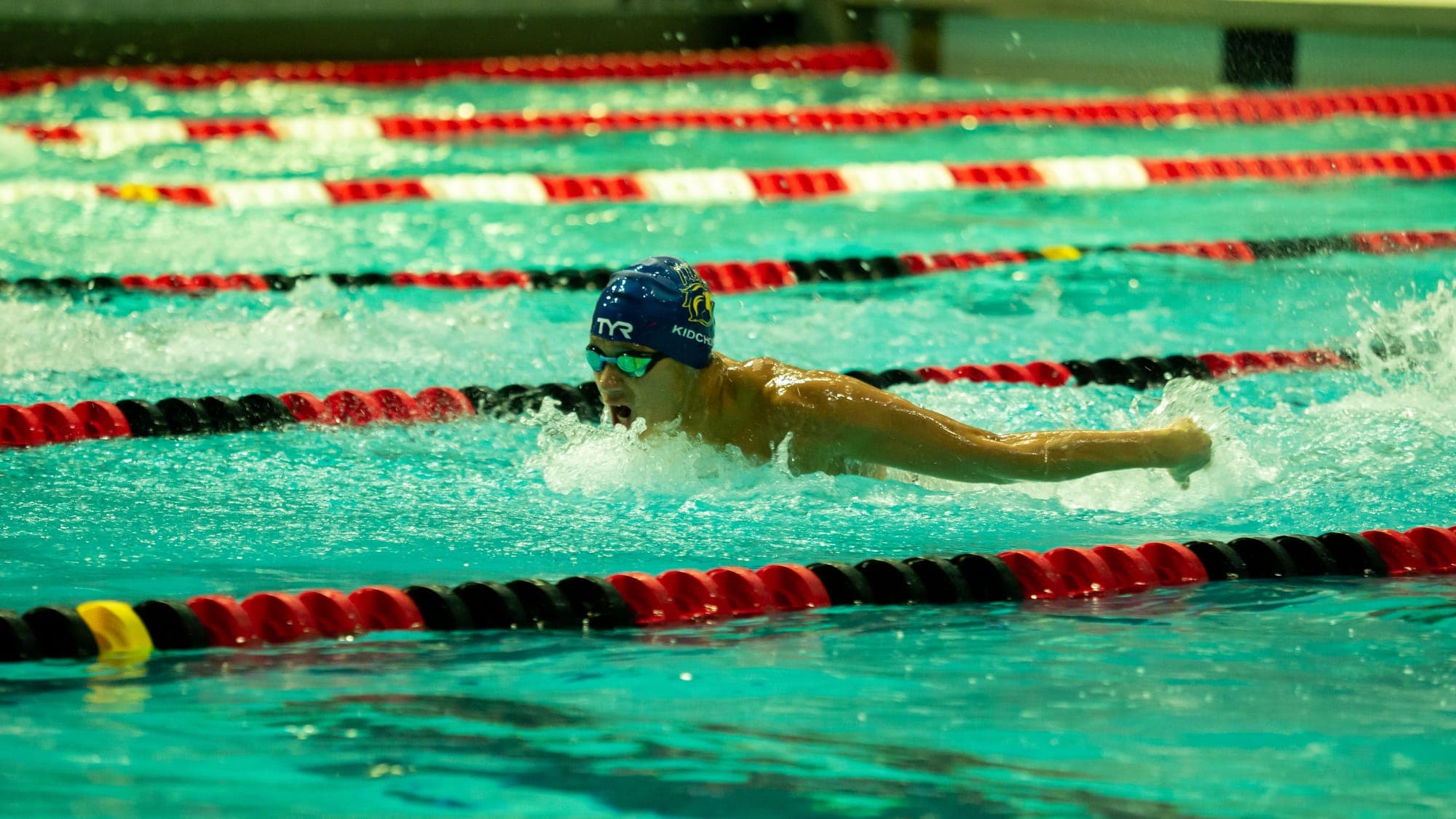 Butterfly swimmer mid-stroke between red lane ropes, water sheeting off his extended arms